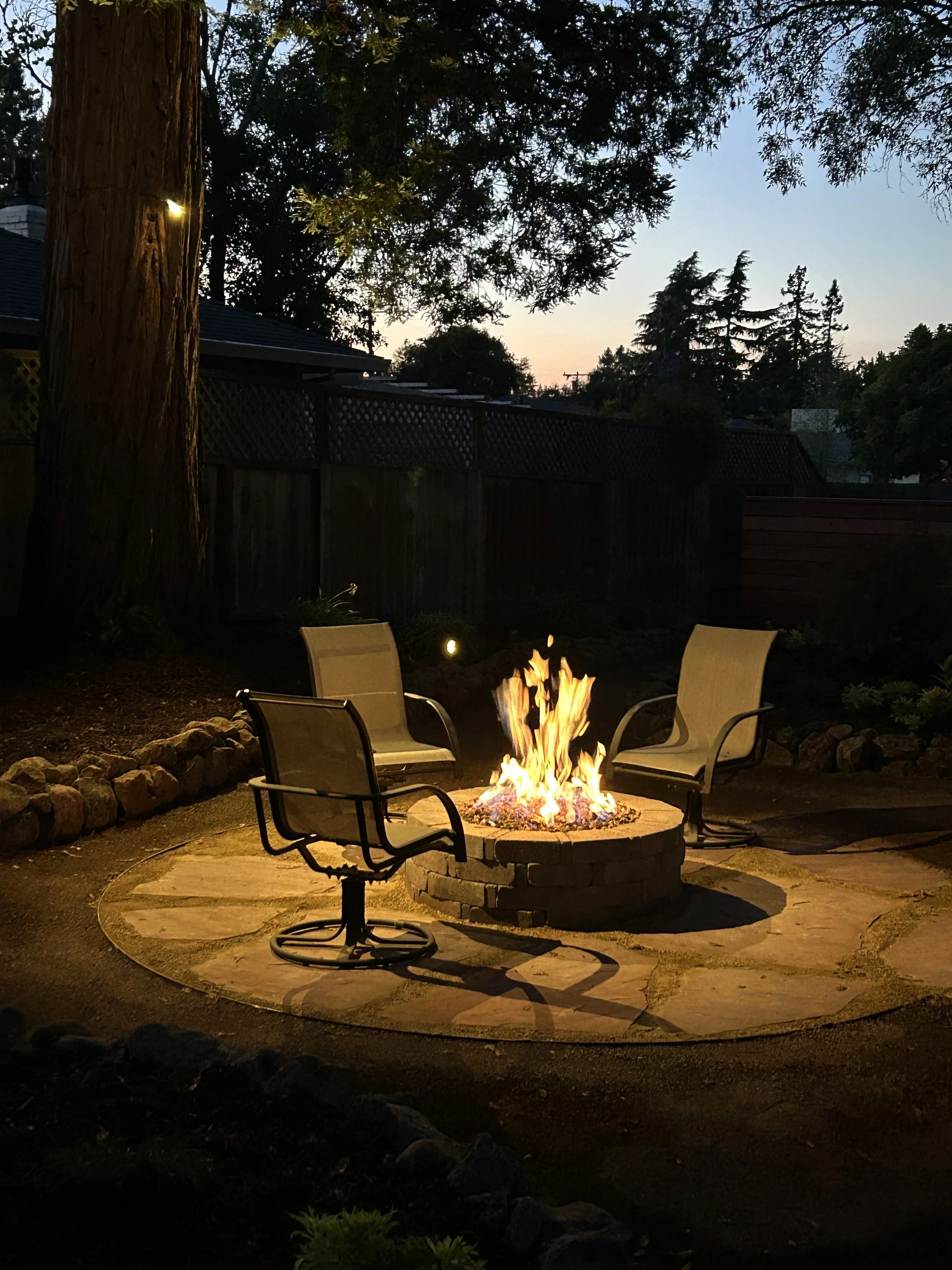 A backyard at dusk featuring a fire pit with flames, surrounded by four chairs, with trees and a fence in the background.