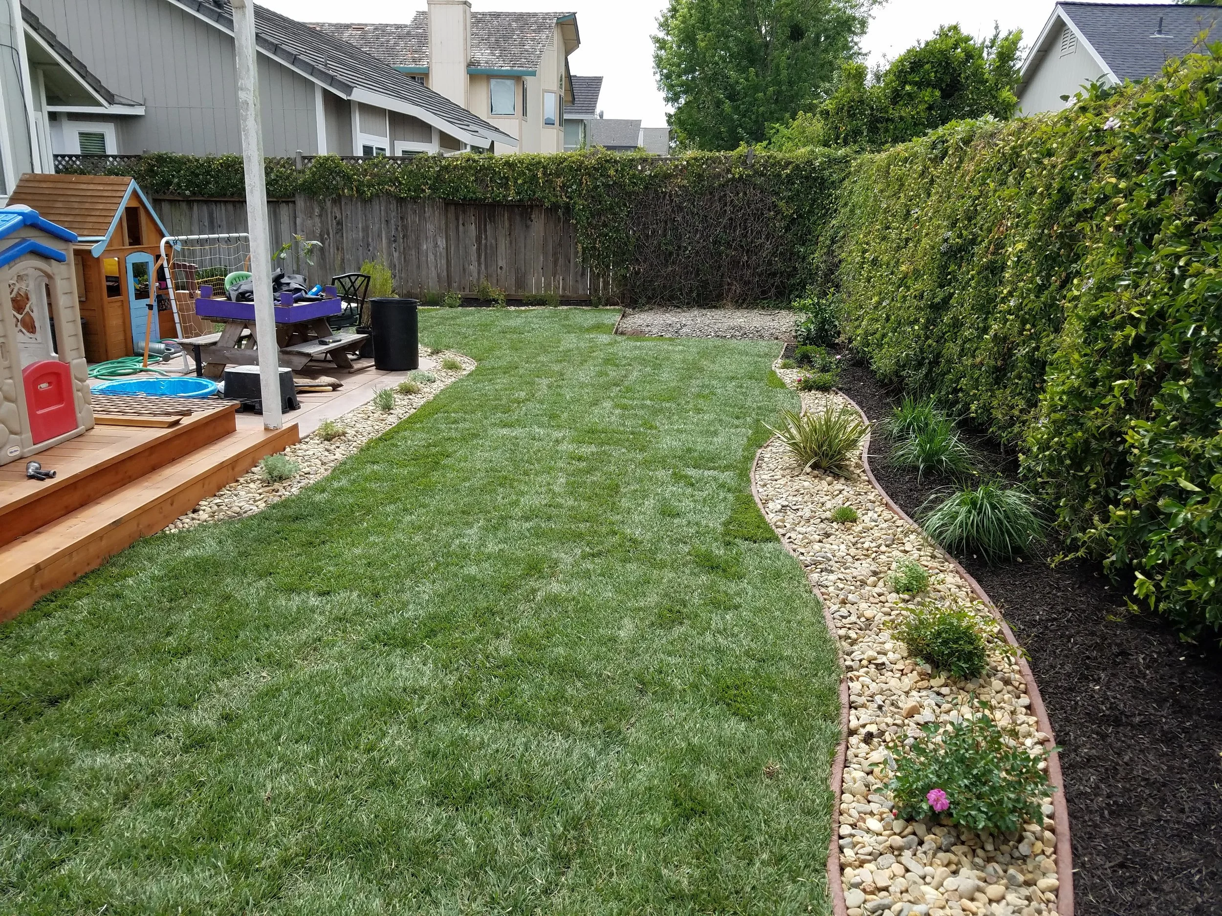 A backyard with a small playground on a wooden deck, green grass, and a garden with shrubs and rocks, surrounded by a wooden fence and neighboring houses and trees in the background.
