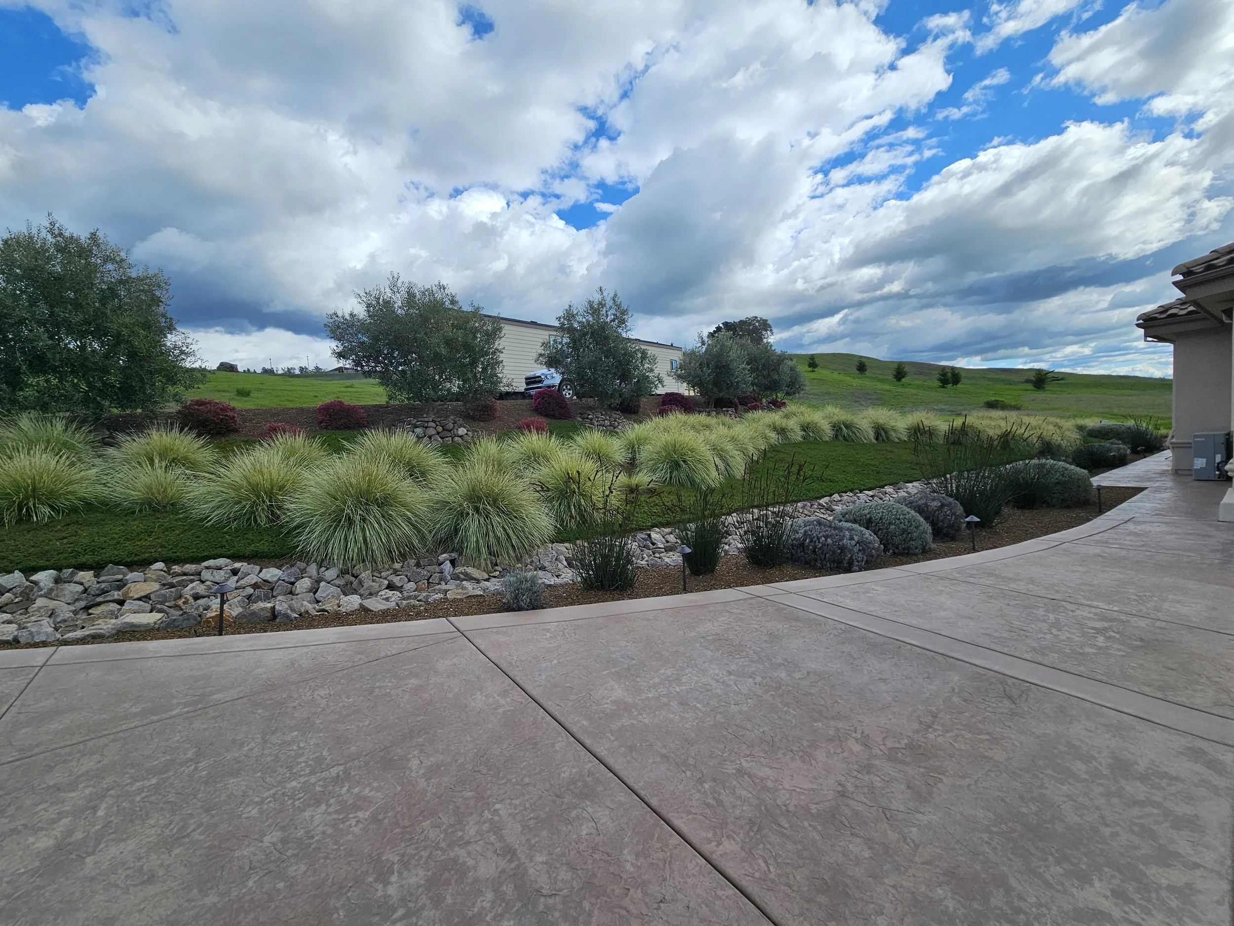 View of a backyard garden with a concrete patio, landscaped with rocks, bushes, and trees. In the background, a mobile home or RV is parked on a grassy hill under a partly cloudy sky.