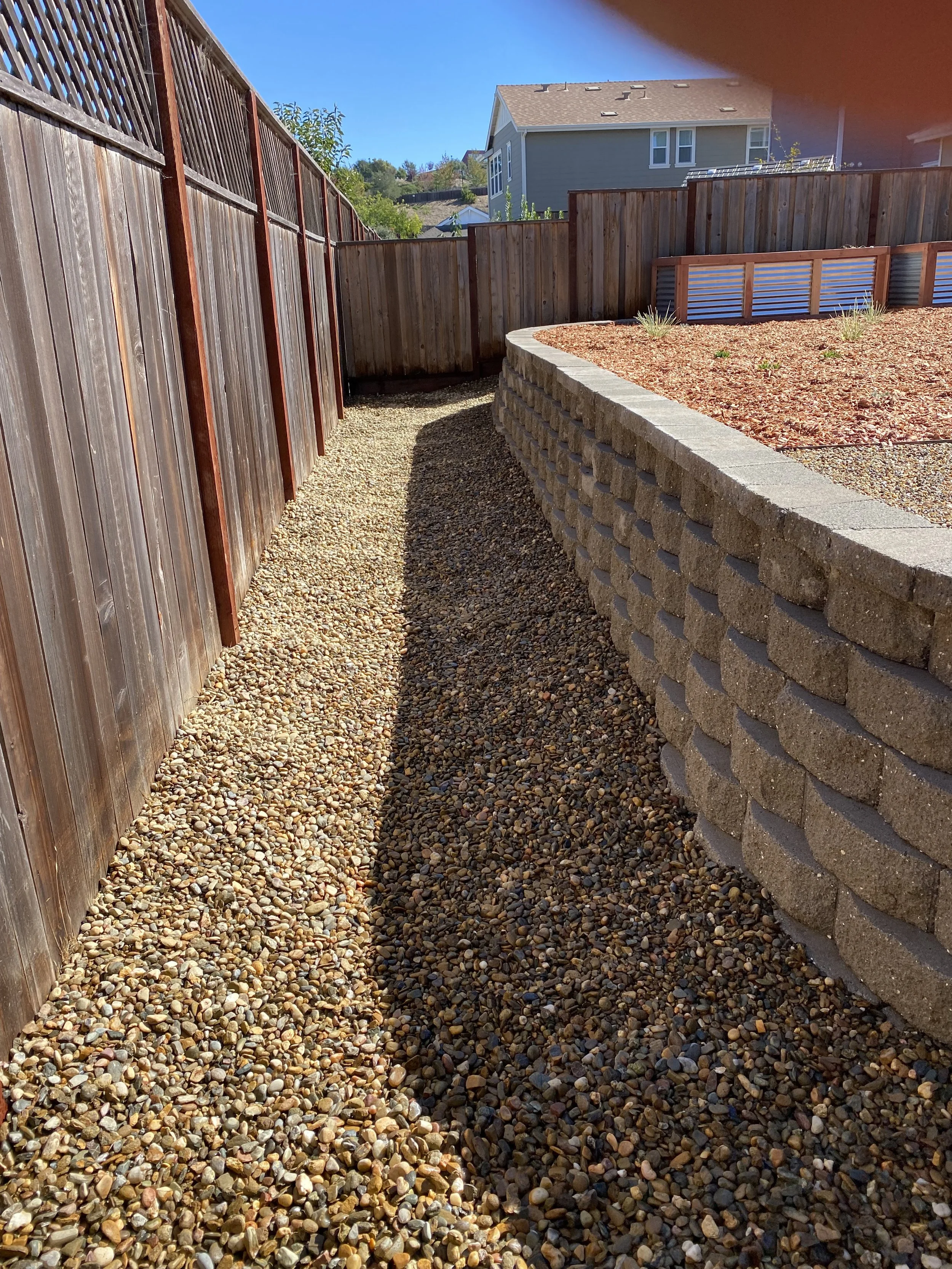 A backyard with a gravel pathway along a wooden fence, with a raised garden bed with plants and a neighboring house in the background under a clear blue sky.