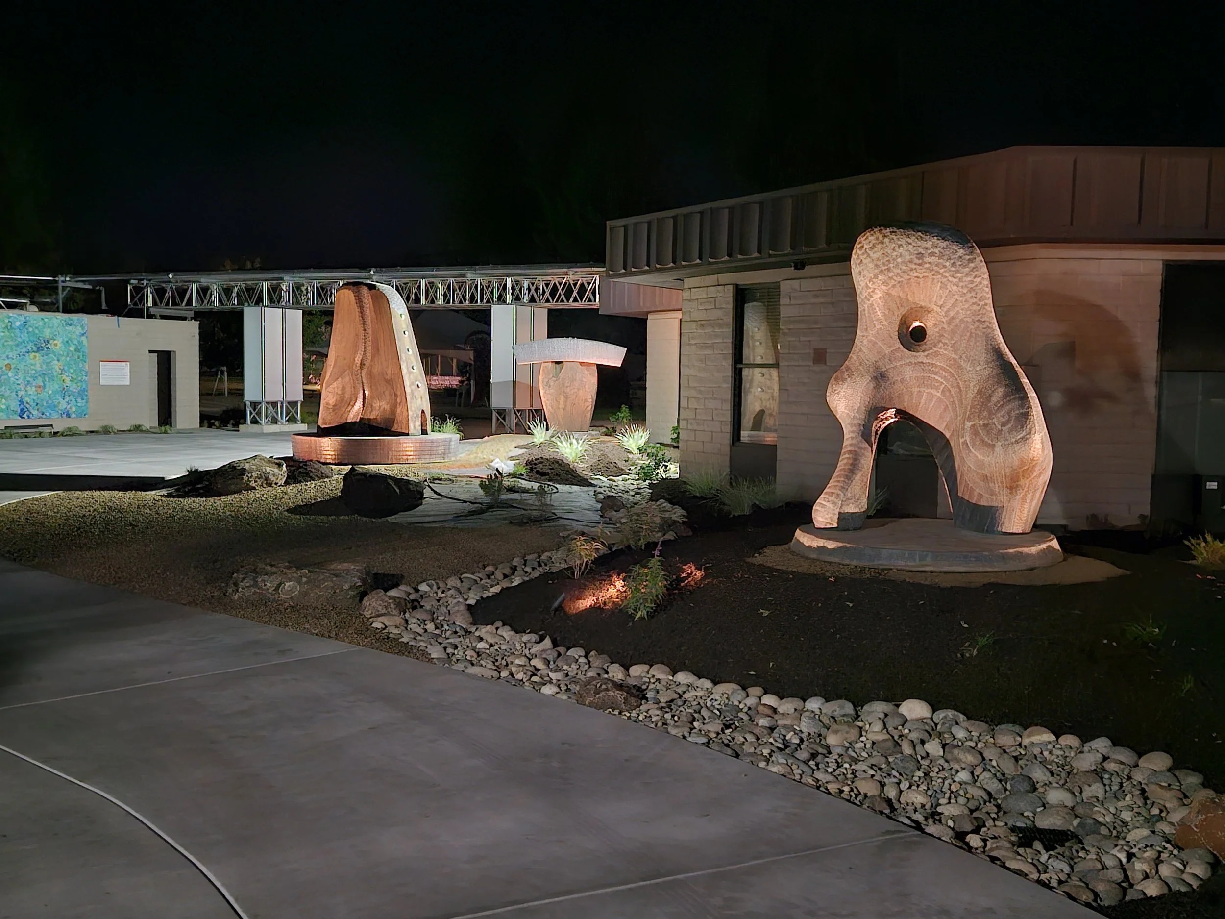 Nighttime exterior view of a modern building with large wooden sculpture of an animal, likely an elephant, illuminated by ground lighting, with architectural elements, rocks, and plants in the foreground.