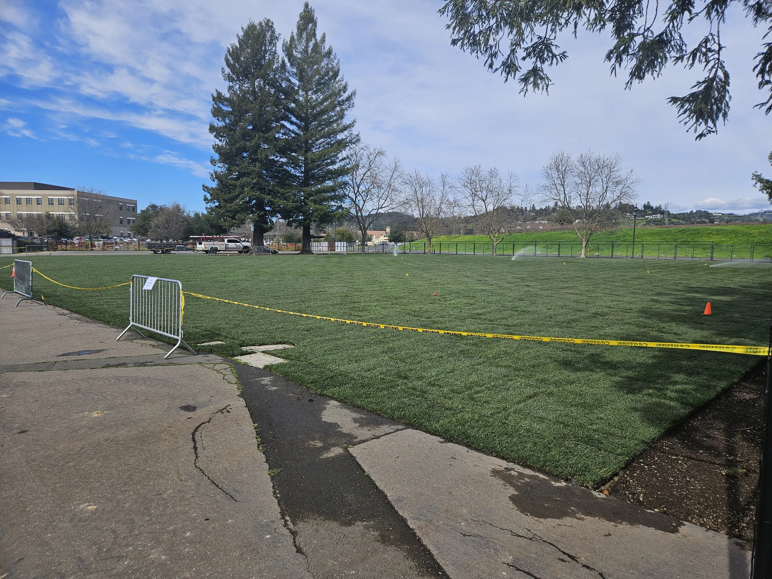 A park with green grass, large trees, and some smaller trees, with a large building in the background under a partly cloudy sky. There are water sprinklers watering the grass, and orange cones and metal barriers near the edge of the grass area.