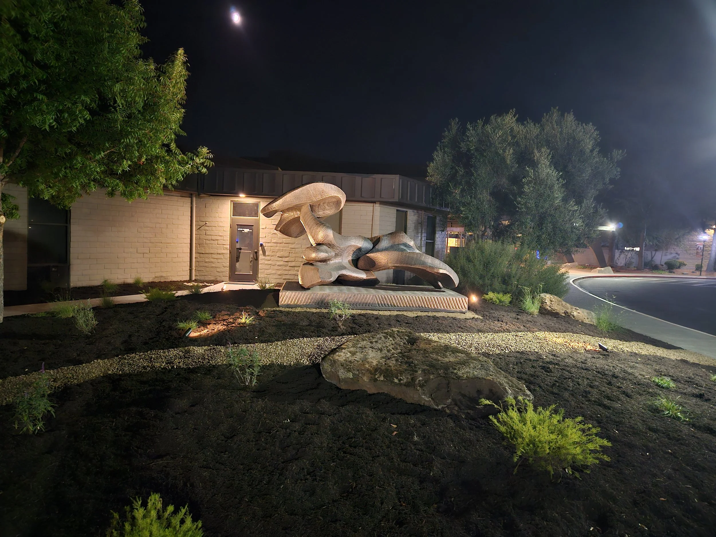 Night scene of an outdoor landscaped area with trees, a modern sculpture, and building with lights, under a moonlit sky.