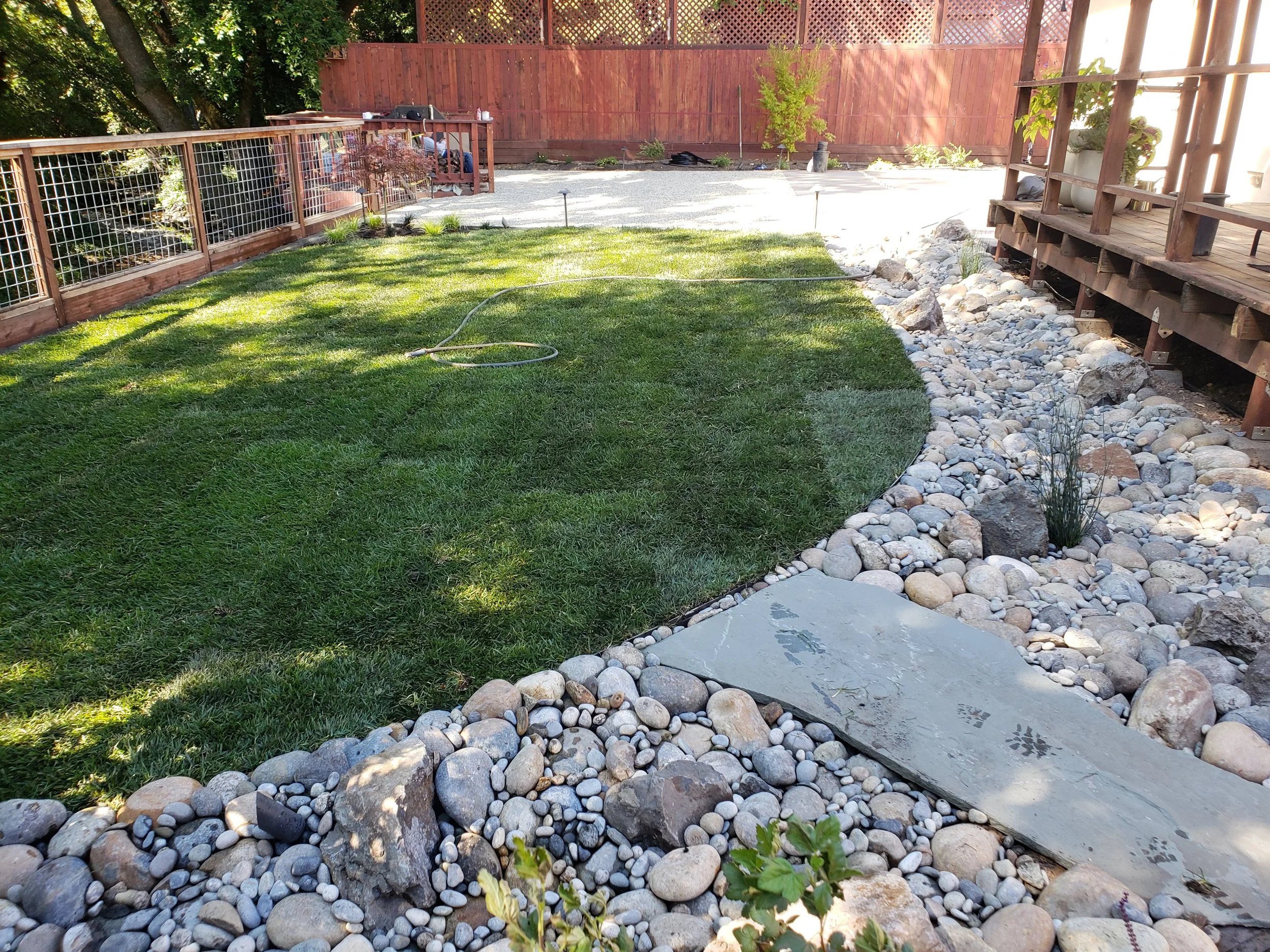 A backyard with a grass lawn, a wooden deck with potted plants, a red wooden fence, and a yard area with rocks and gravel.