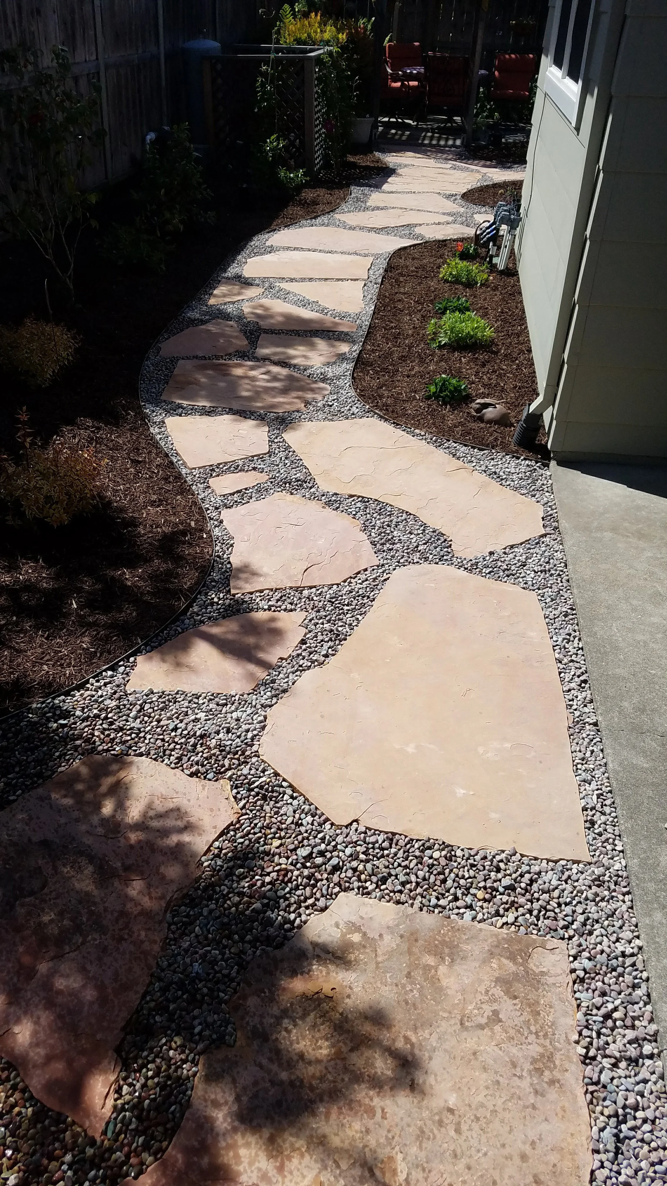 A winding stone pathway made of large flat stones bordered by small pebbles, leading through a garden area with plants and a patio with chairs in the background.