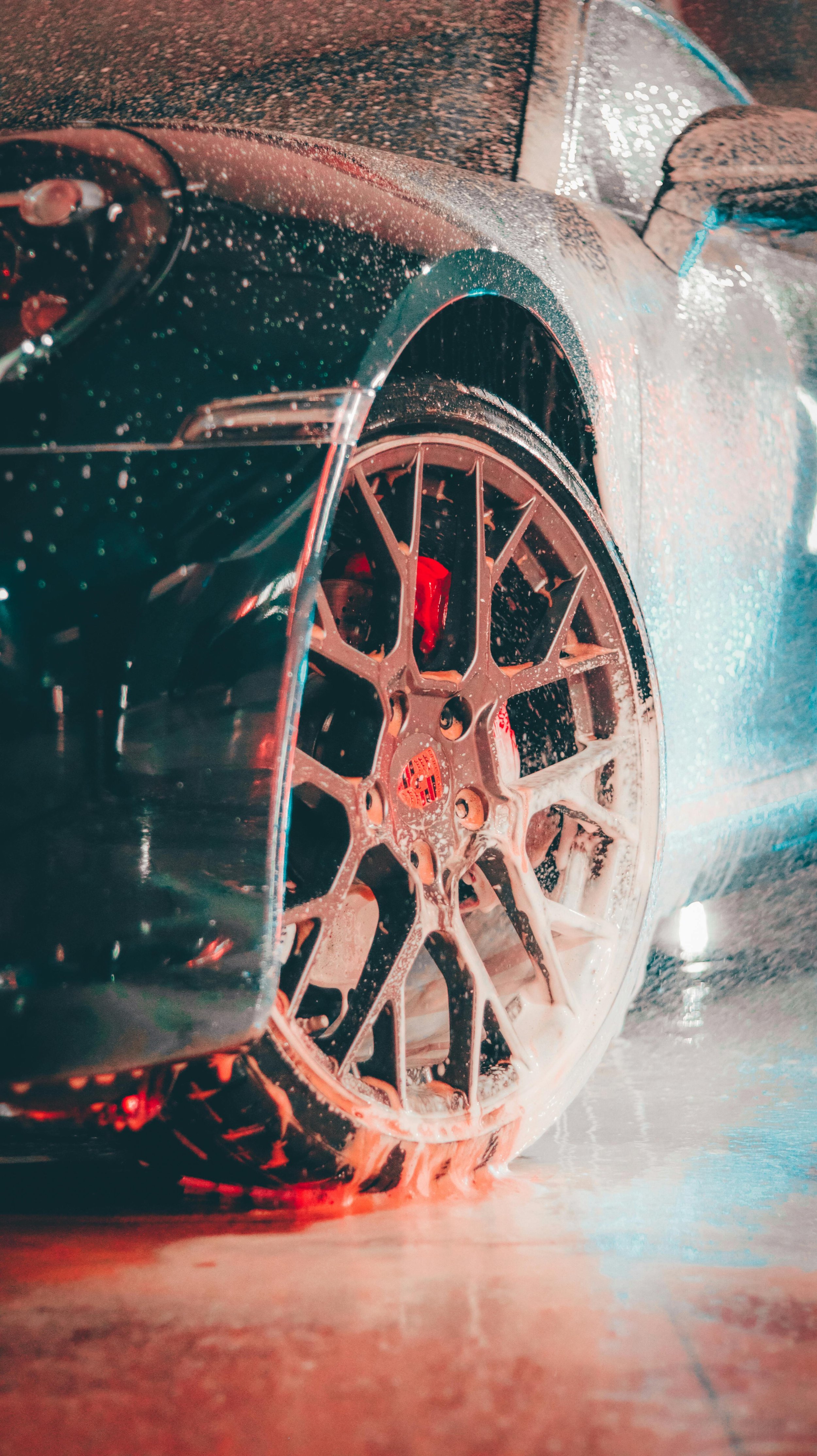 Close-up of a sports car with a wet surface during car wash, focusing on the front wheel and tire with foam splashing around it.
