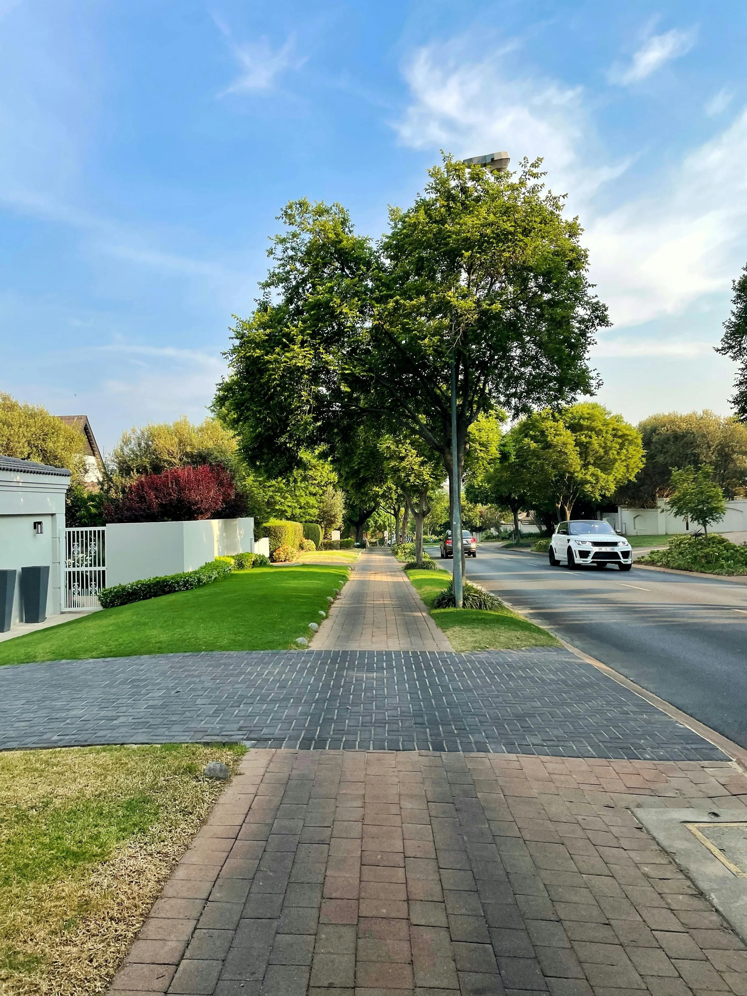 A suburban street scene showing a paved sidewalk lined with trees, a grassy lawn on the left, and a street with moving cars on the right, under a partly cloudy sky.
