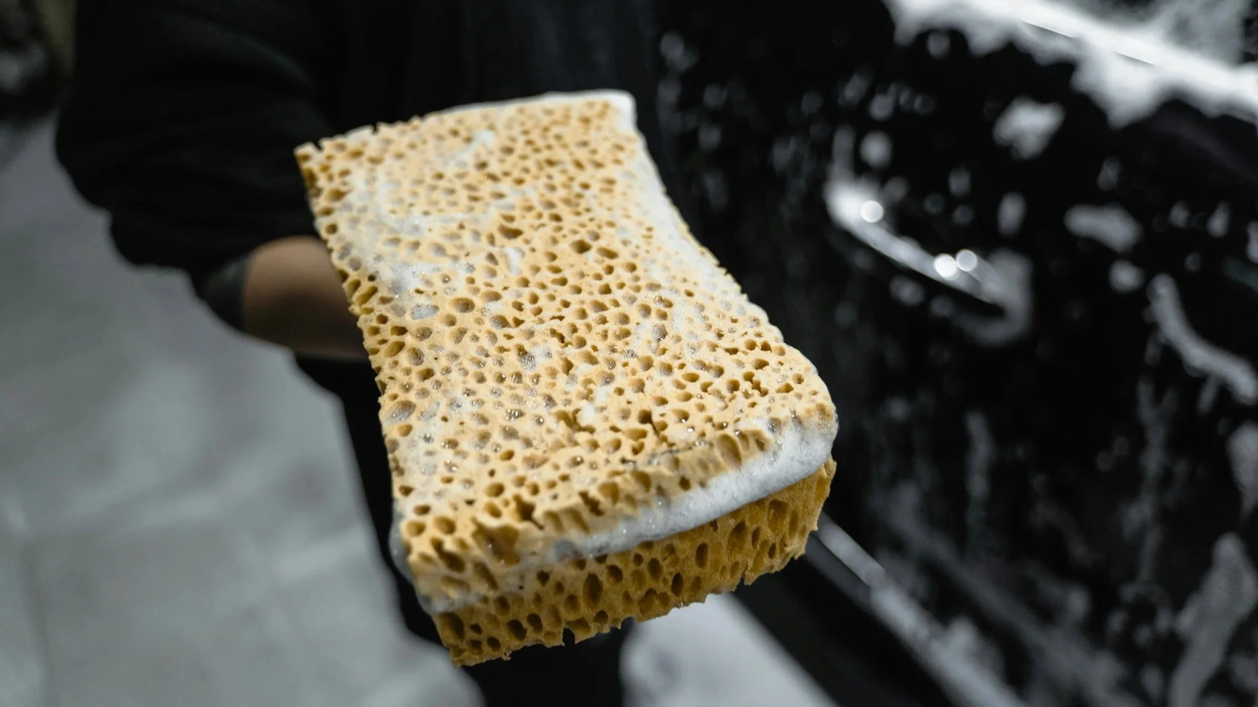 Close-up of a sponge scrubber with foam, held by a car and motorcycle detailer wearing black gloves, with a black car in the background.