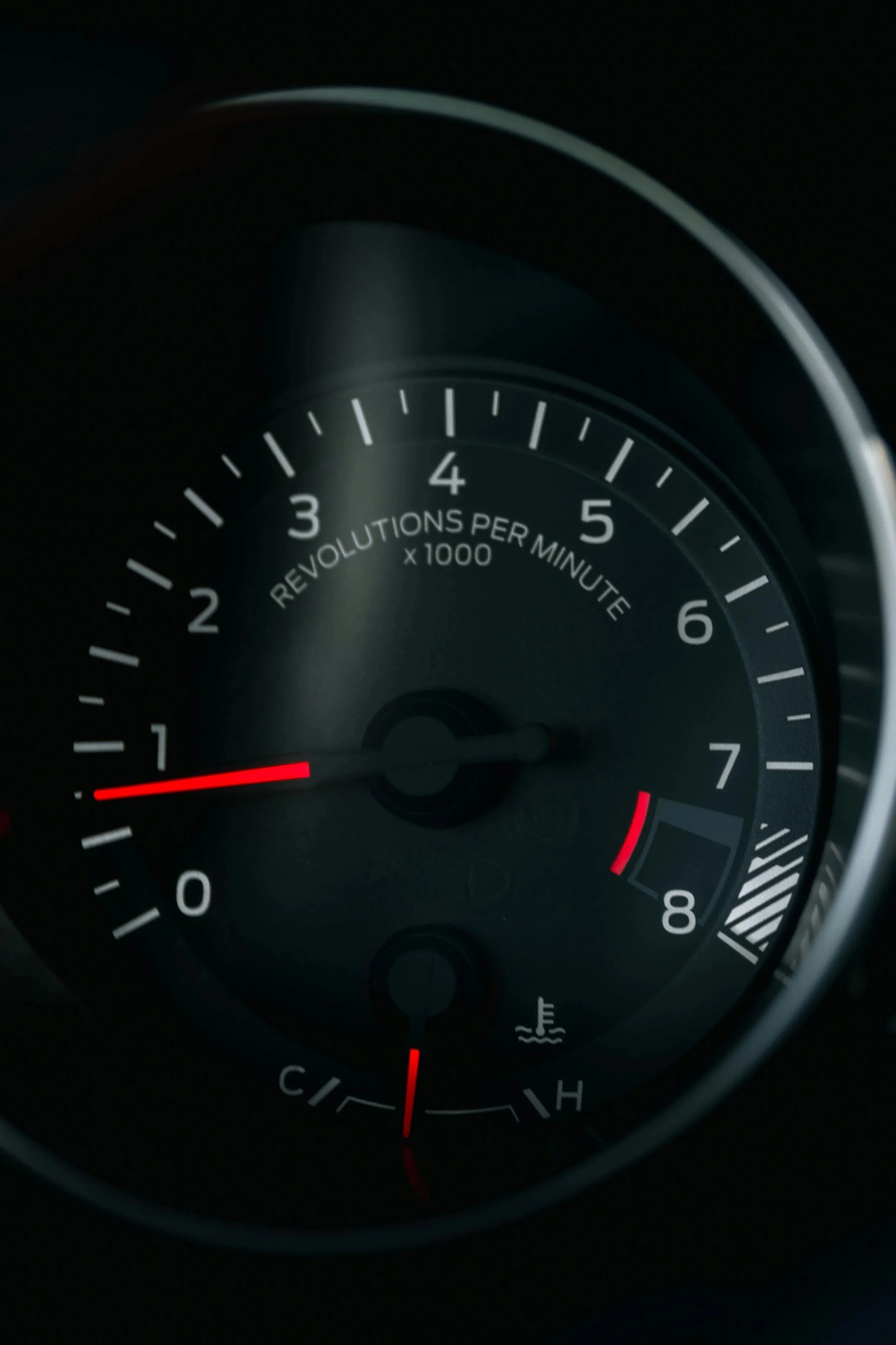 Car dashboard tachometer gauge with needle pointing to 1, indicating 1000 revolutions per minute, and a temperature gauge below showing a low reading.