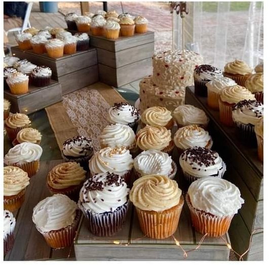 Assorted cupcakes on a table, some with white, chocolate, or caramel frosting, and decorated with sprinkles or chocolate shavings, with larger cupcakes and a cake in the background.