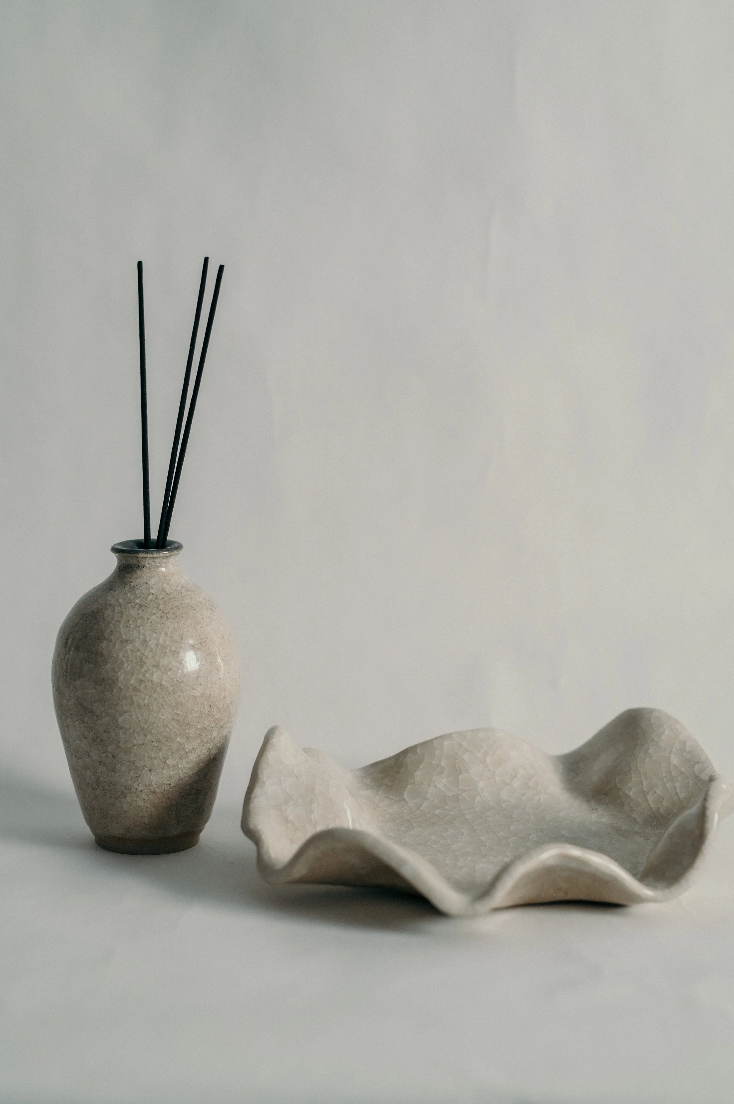 A beige textured ceramic vase with three black reed sticks and a beige wavy ceramic dish on a white surface against a plain light background.