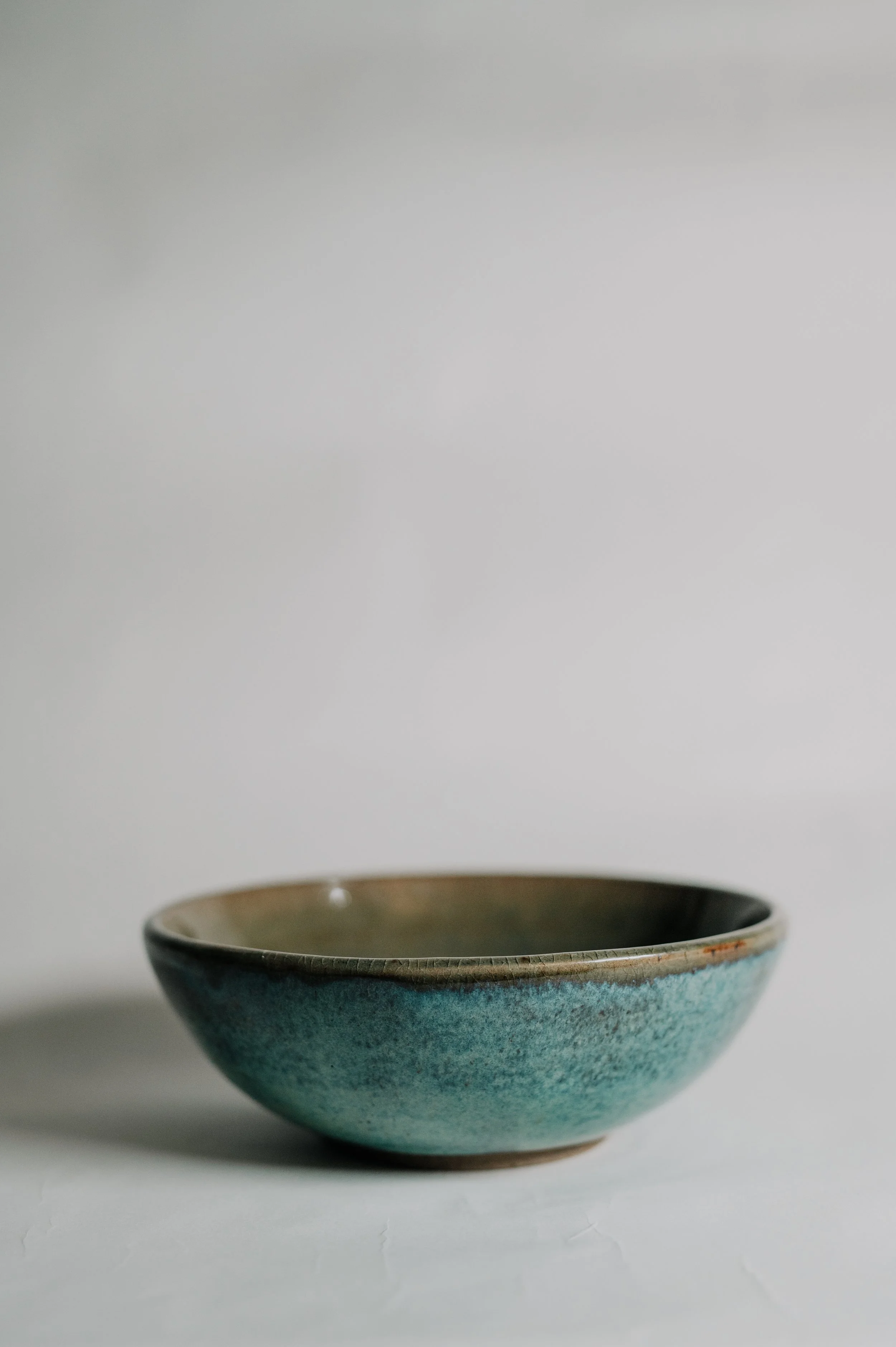 A ceramic bowl with a brown rim and teal glaze, placed on a white surface in front of a plain white background.