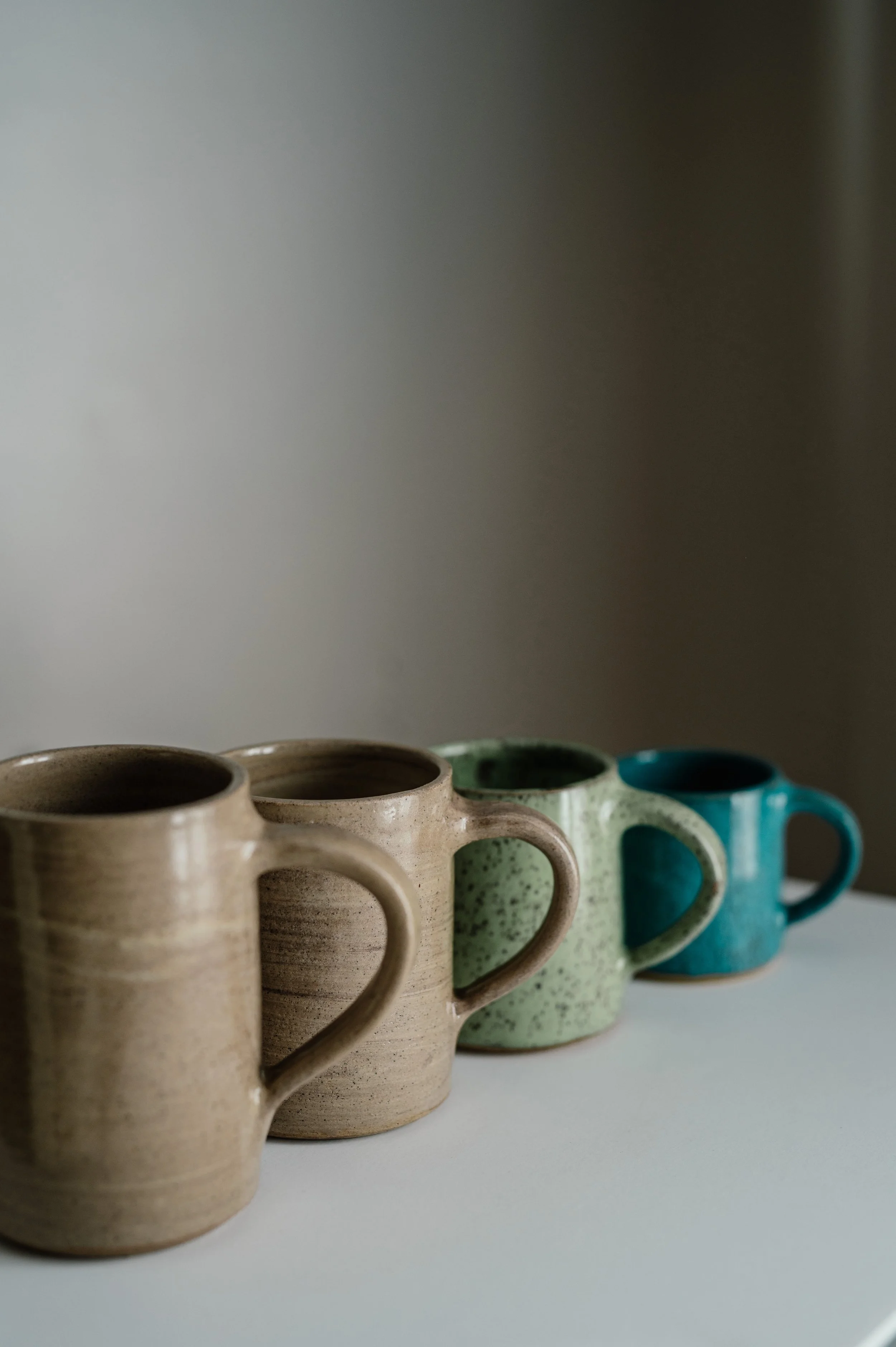 Four ceramic mugs in beige, green, and blue hues lined up on a white surface against a plain background.