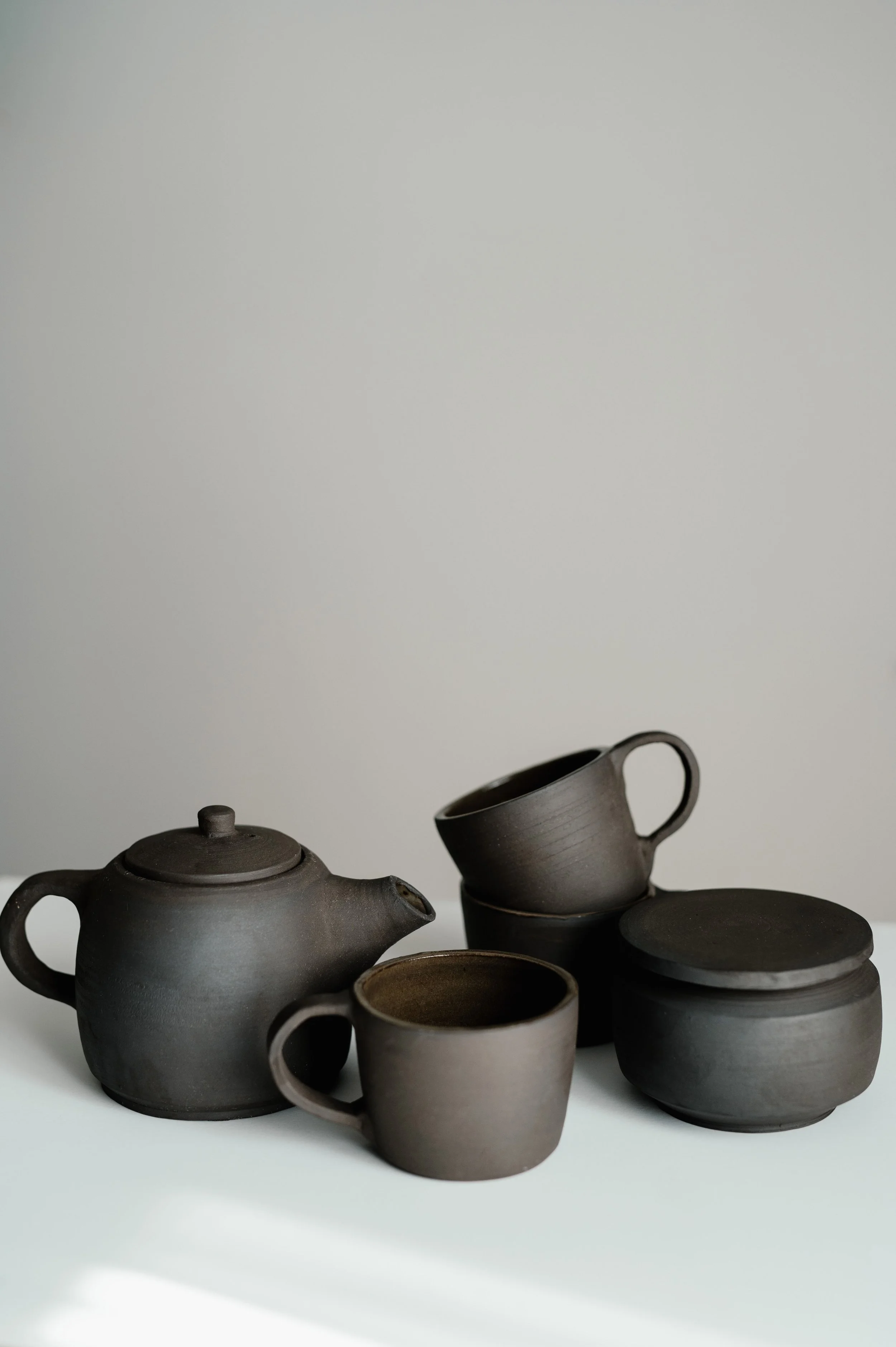 Set of dark gray ceramic teapot, cups, and containers on white surface with plain gray background.
