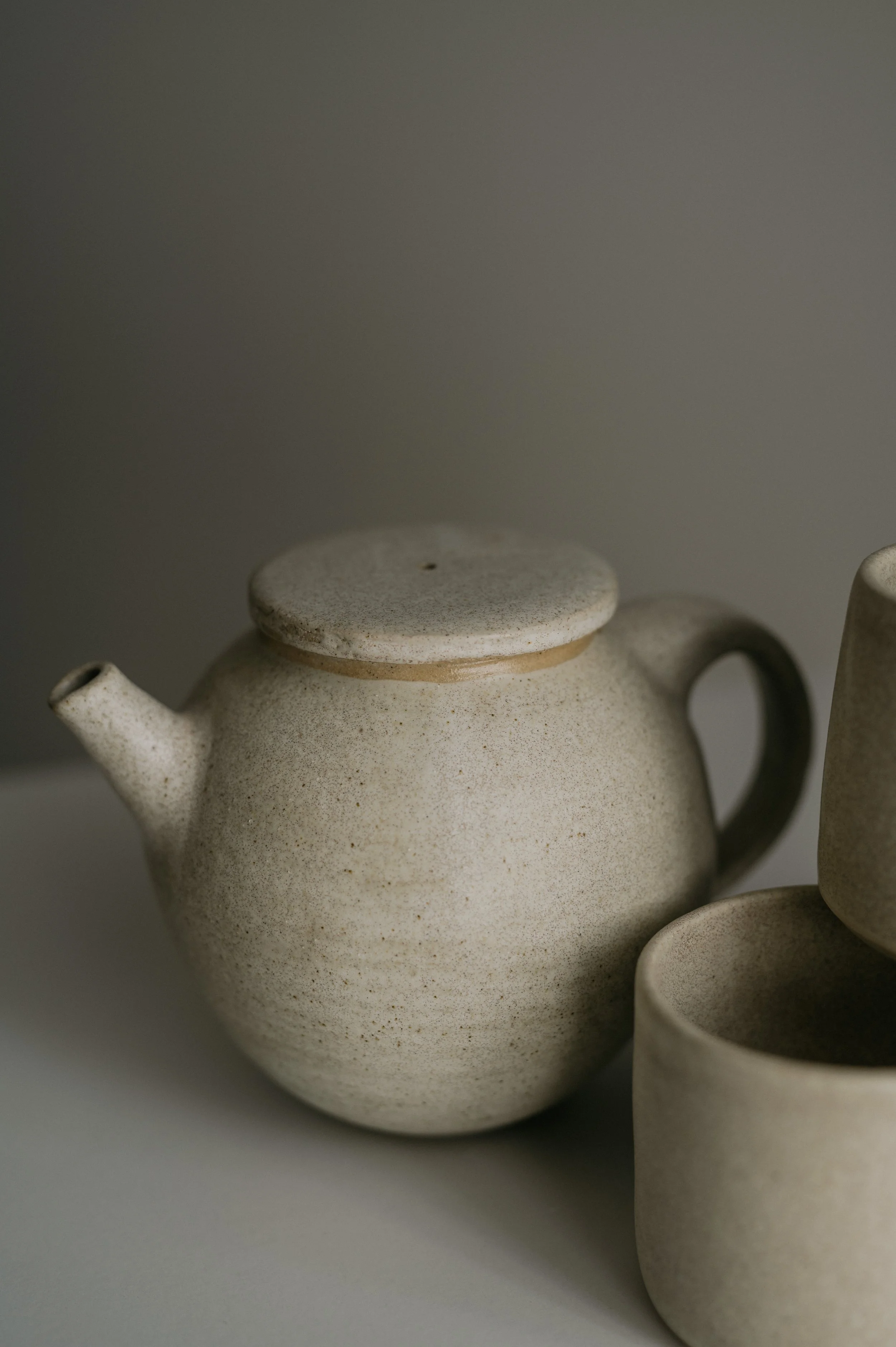 Cream-colored ceramic teapot with a handle and a lid, alongside matching cups, on a plain surface.