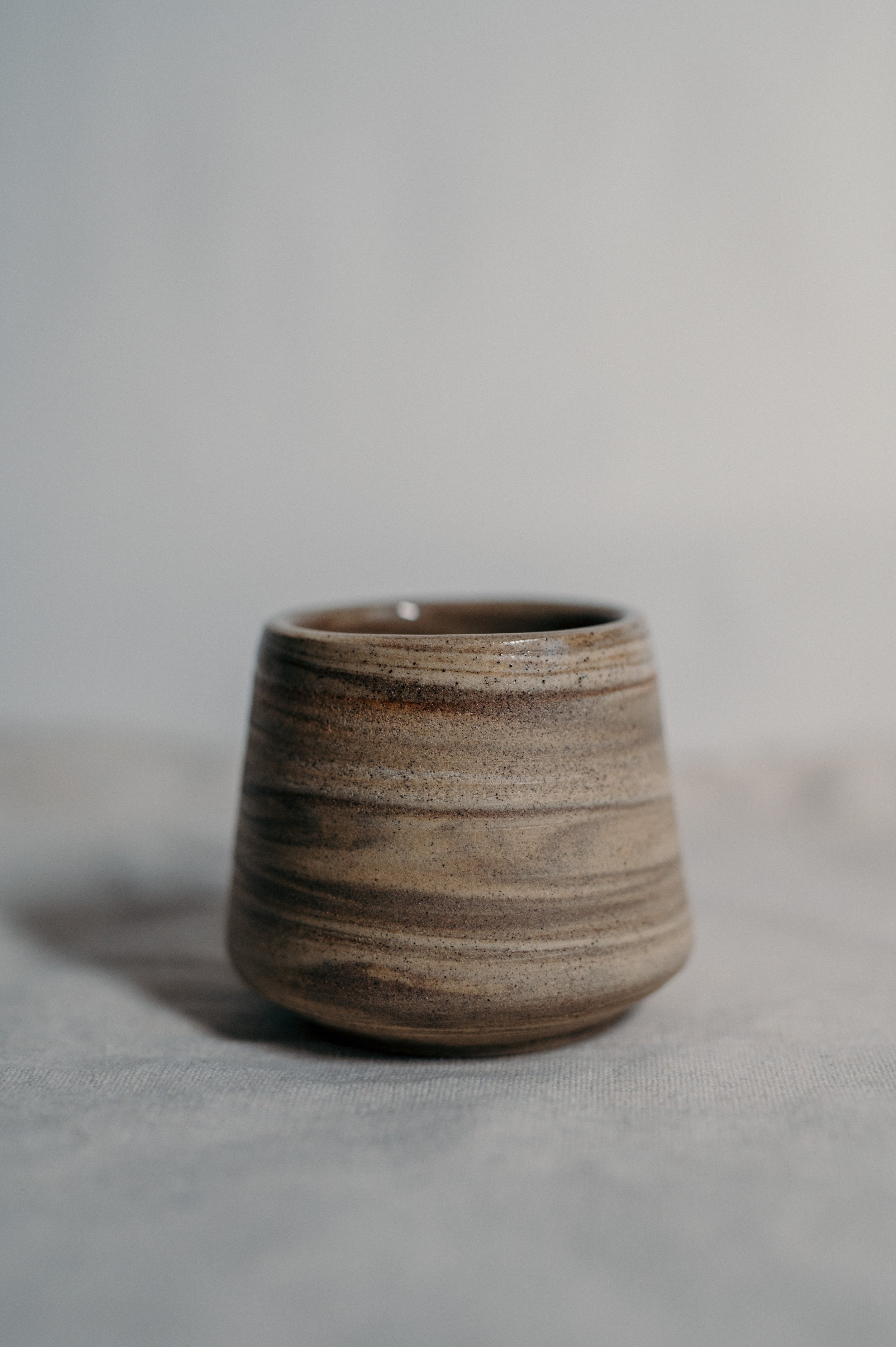 A small, round, ceramic cup with a striped, earthy color pattern on a light fabric surface, against a plain light background.