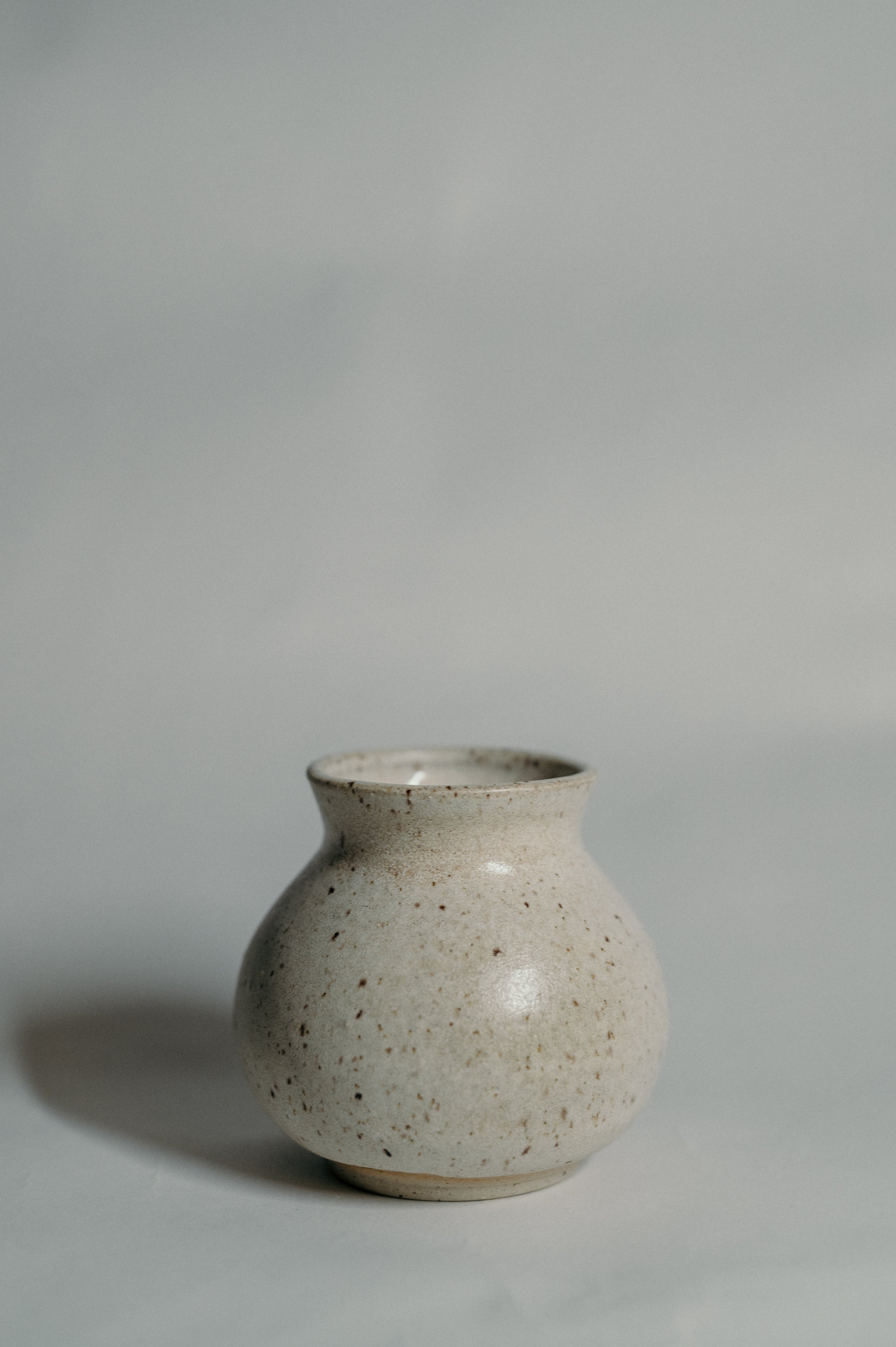 A small, ceramic vase with a speckled, off-white glaze, set against a plain, light gray background.