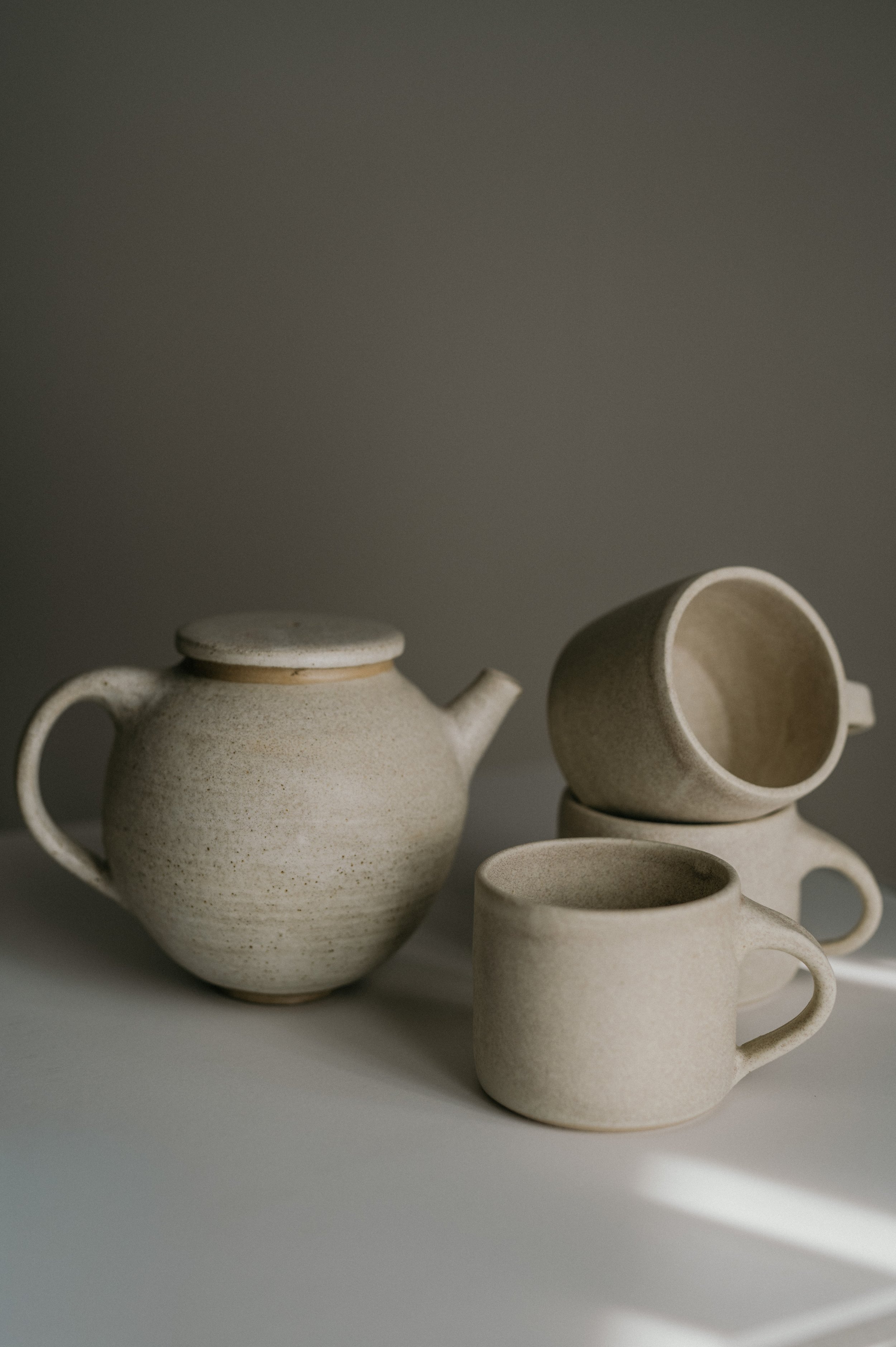 Cream-colored ceramic teapot and three cups on a white surface with shadows and a gray background.