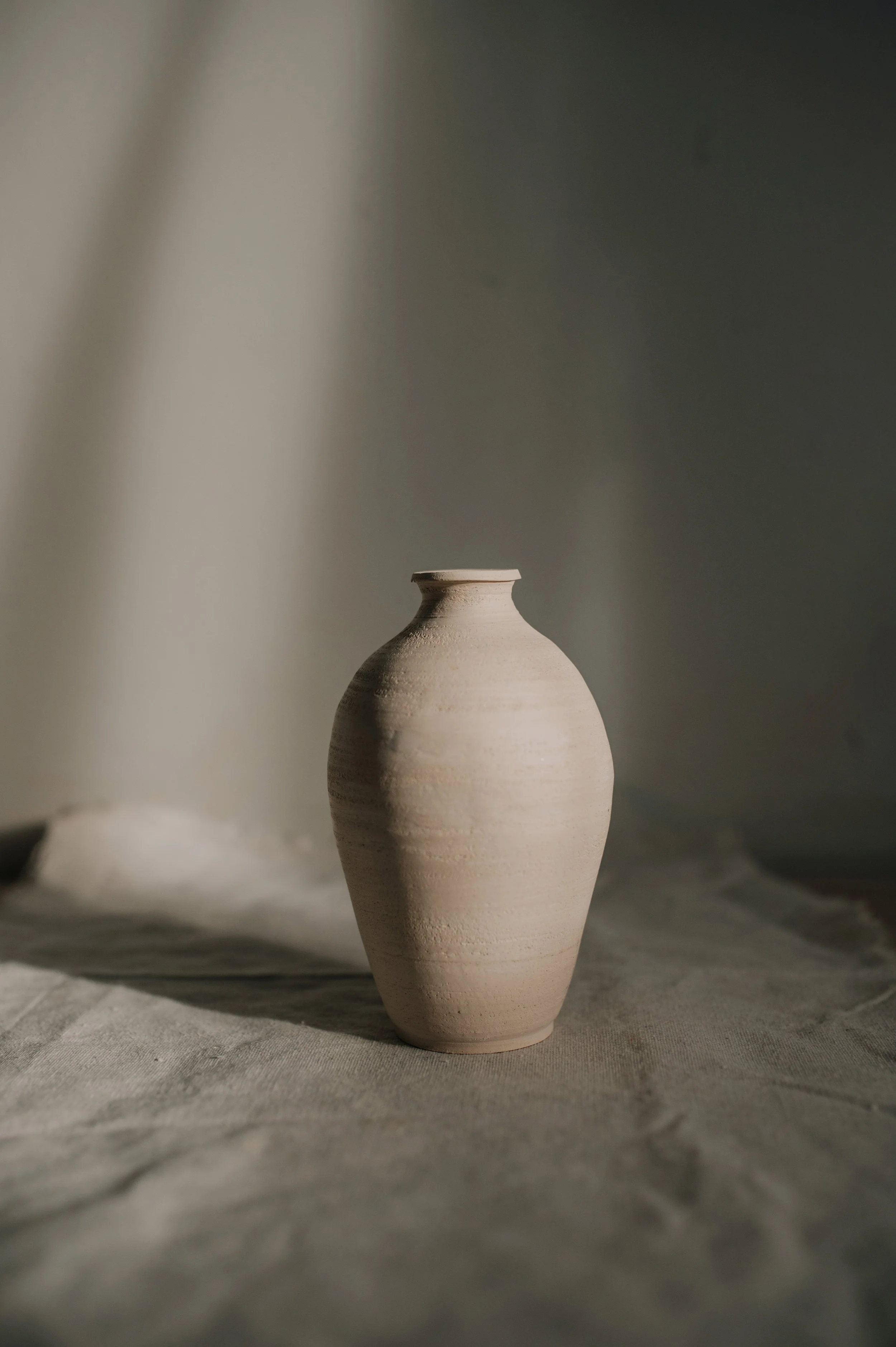 A light-colored ceramic vase with a narrow neck and round body sitting on a textured fabric surface with shadow and light play.