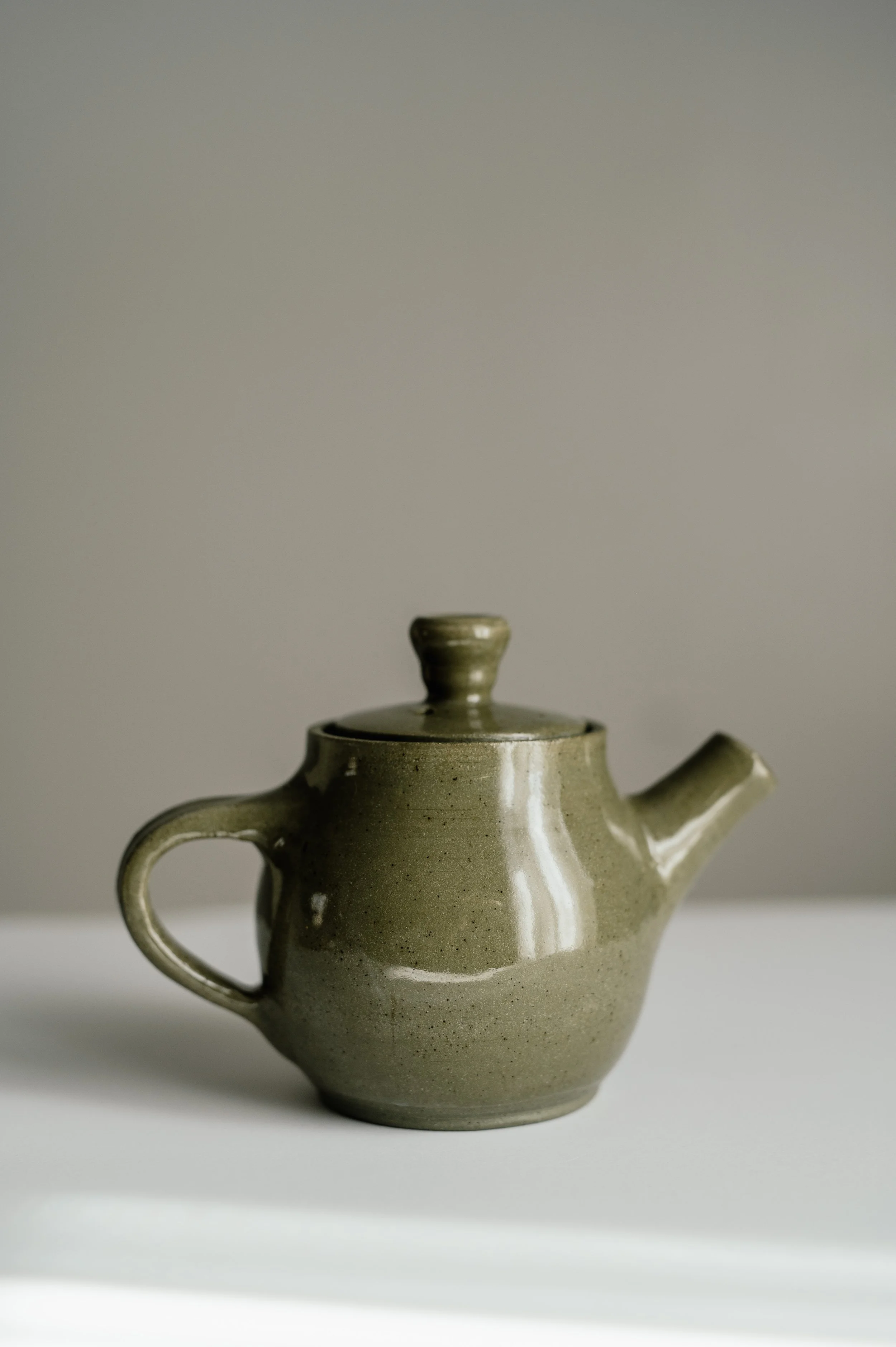 A beige ceramic teapot with a round handle and a short spout, placed on a white surface against a plain light gray background.