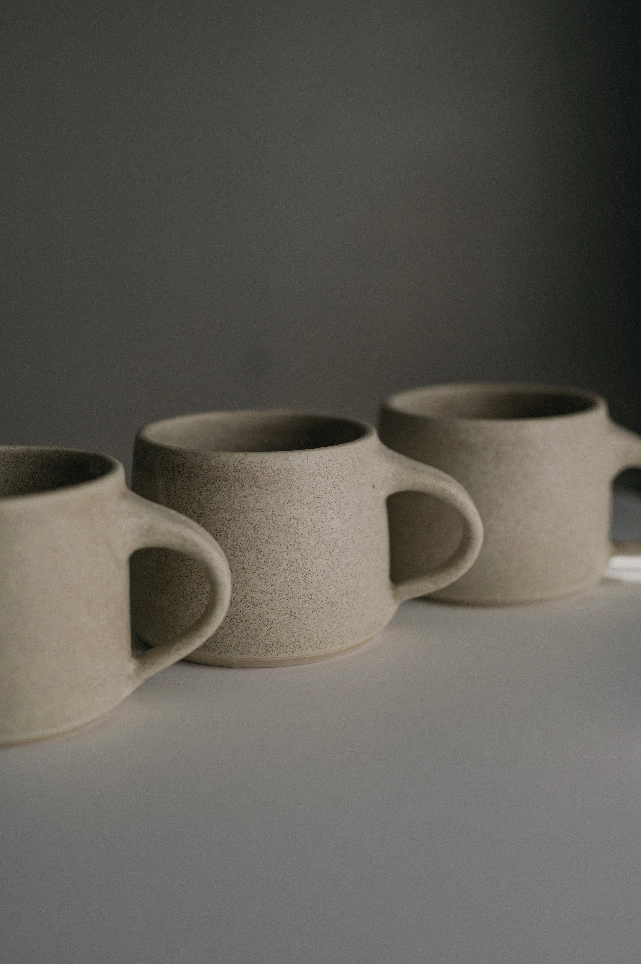 Three beige ceramic mugs with handles placed in a row on a light surface, against a dark background.