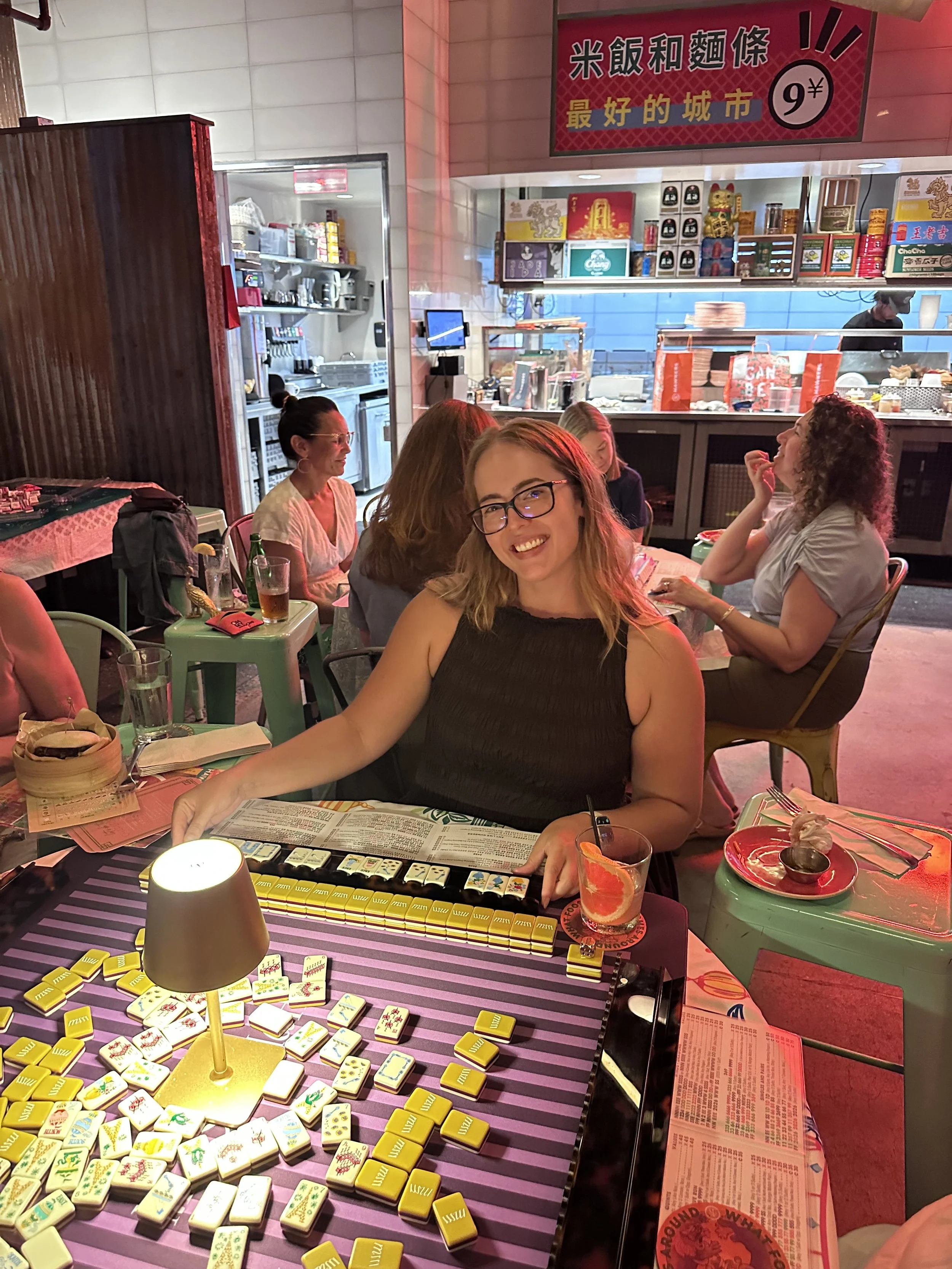 Group of women playing Mahjong at a restaurant table with a game and drinks, with a food counter and colorful signs in the background.