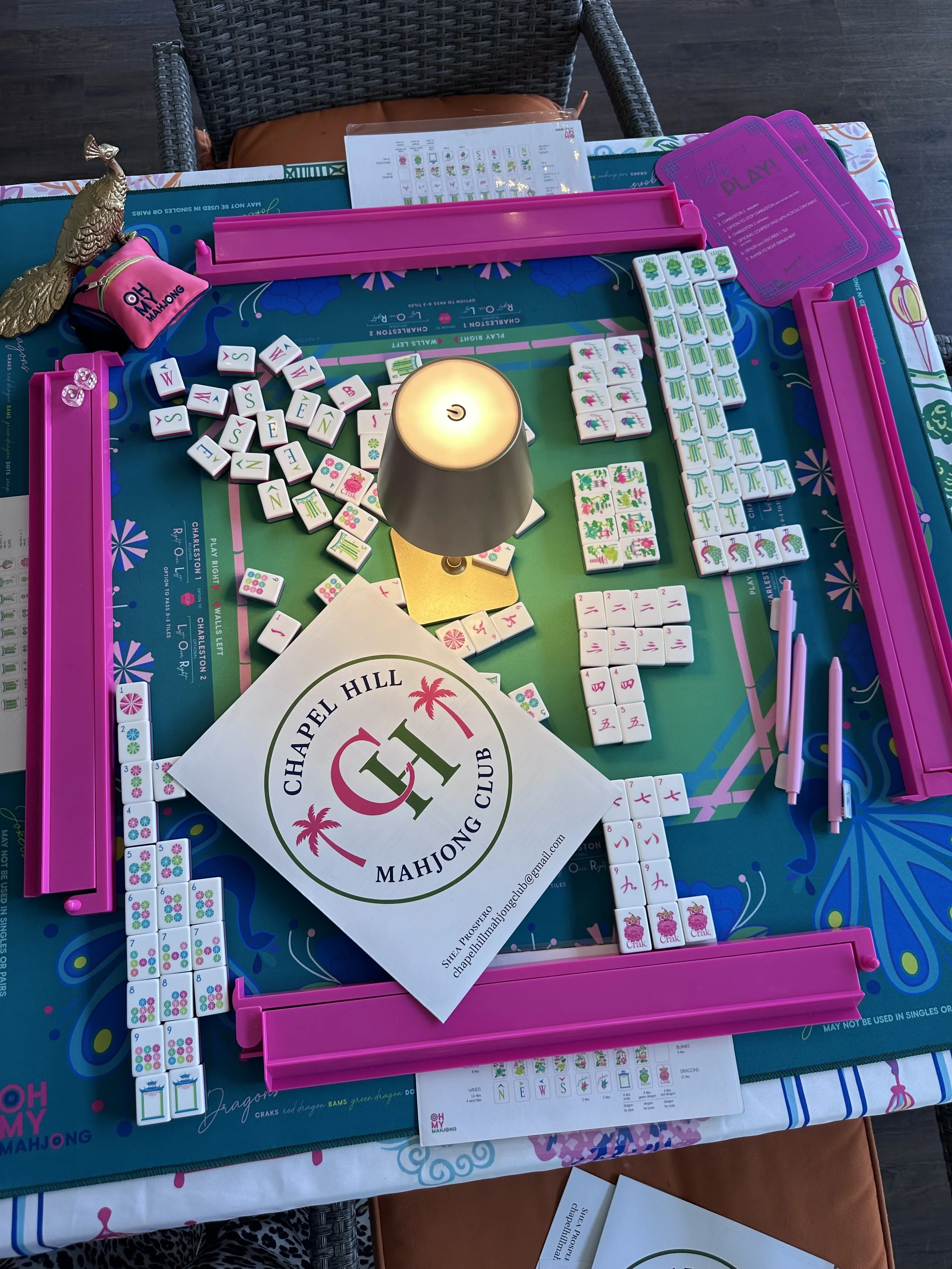 A Mahjong game table with tiles, a pink rack, and accessories, including a candle and a sign from Chapel Hill Mahjong Club.