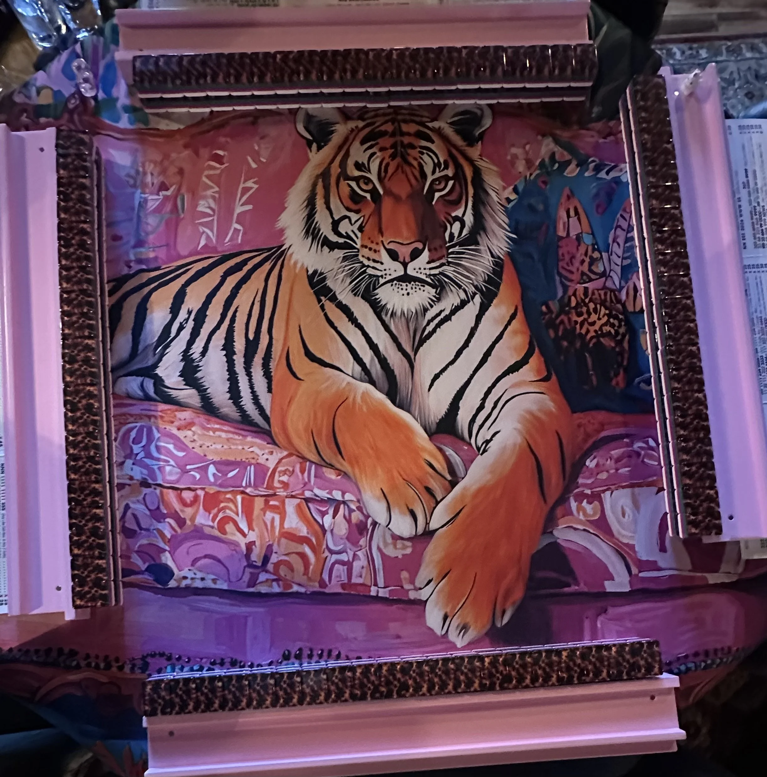 A tiger painting in a pink frame, displayed on a board with a patterned border. Surrounding the frame are various supplies, including pens and paper, indicating it is part of an art display or project.