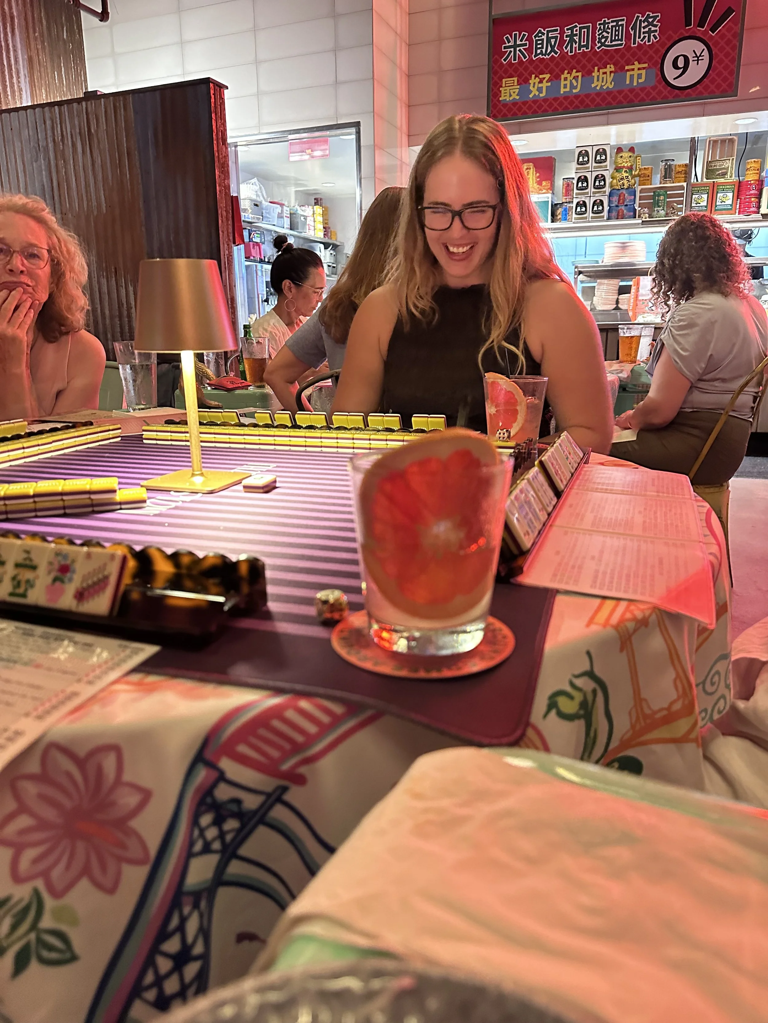 A group of women sitting around a table playing a game at a restaurant. The table has a striped pink and purple tablecloth, and there is a glass of drink with a slice of fruit in the foreground. The restaurant has Asian signage and decor.