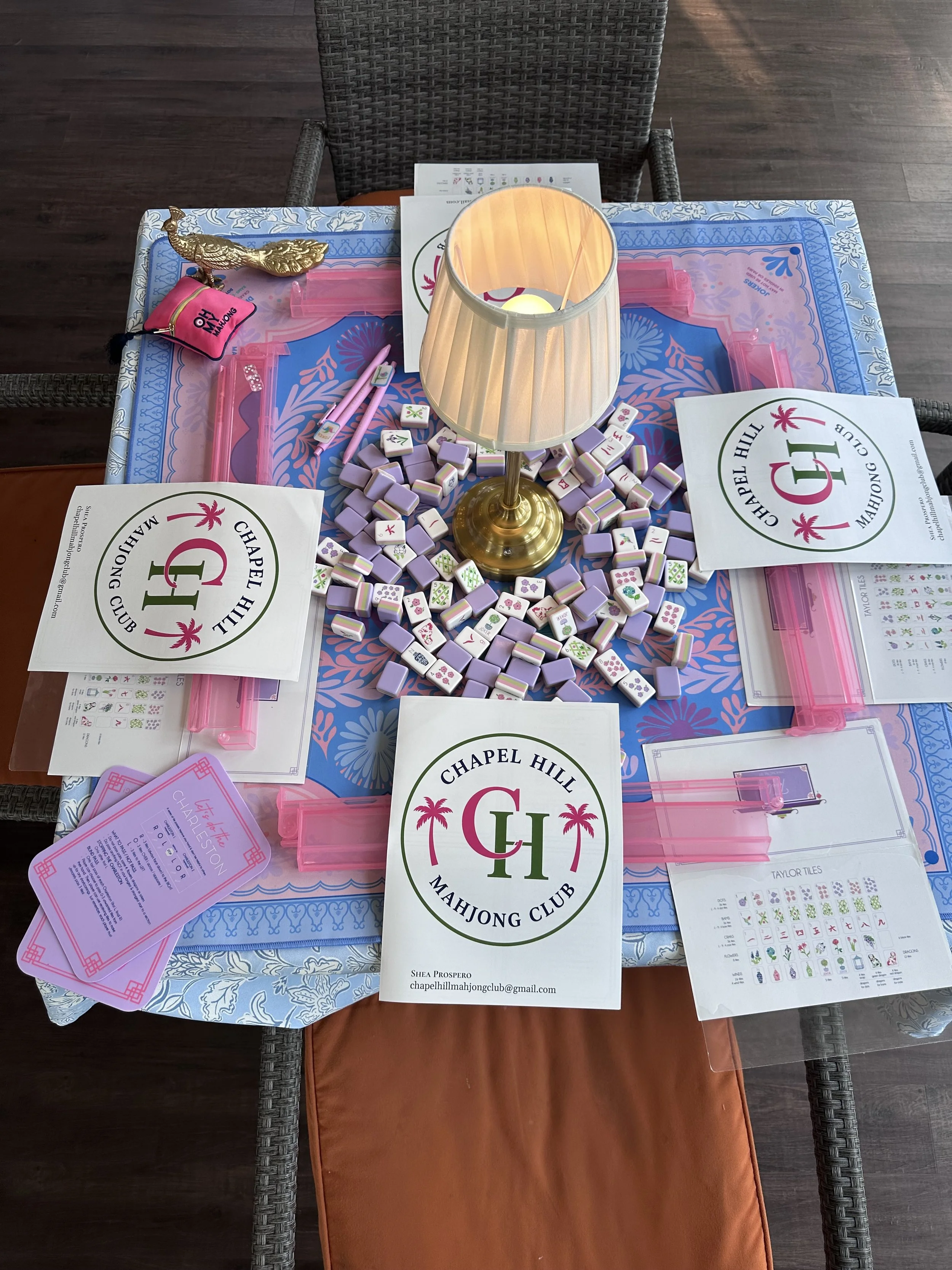 Table with MahJong game pieces, instructions, and a lamp, with a wicker chair behind it. The table surface has a decorative tablecloth, and there are pink plastic boxes for MahJong tiles. The scene is set indoors or on a porch.