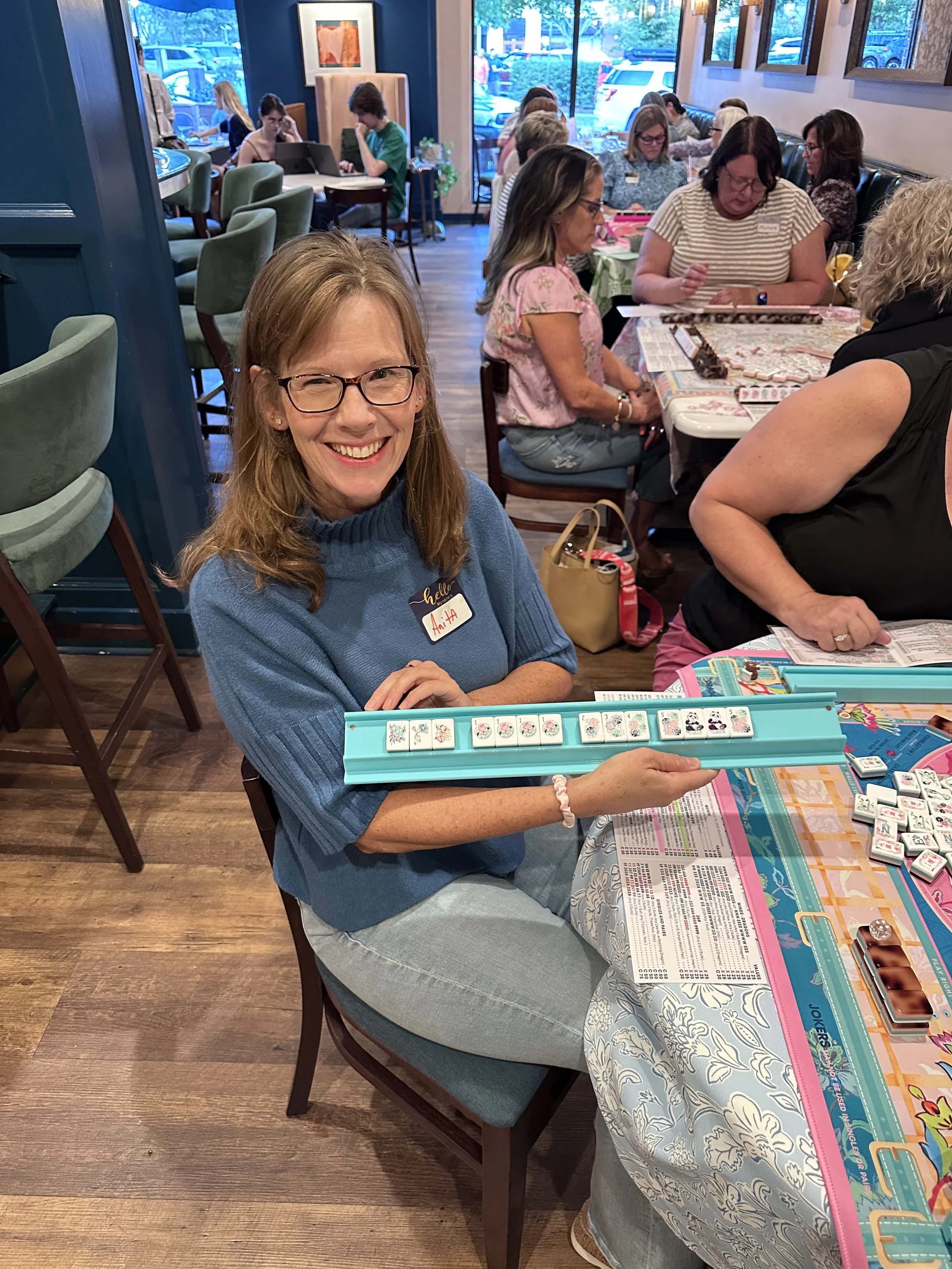 A woman with glasses smiling and holding a Mahjong tile set in a restaurant or cafe setting, with other women engaged in various activities at tables.