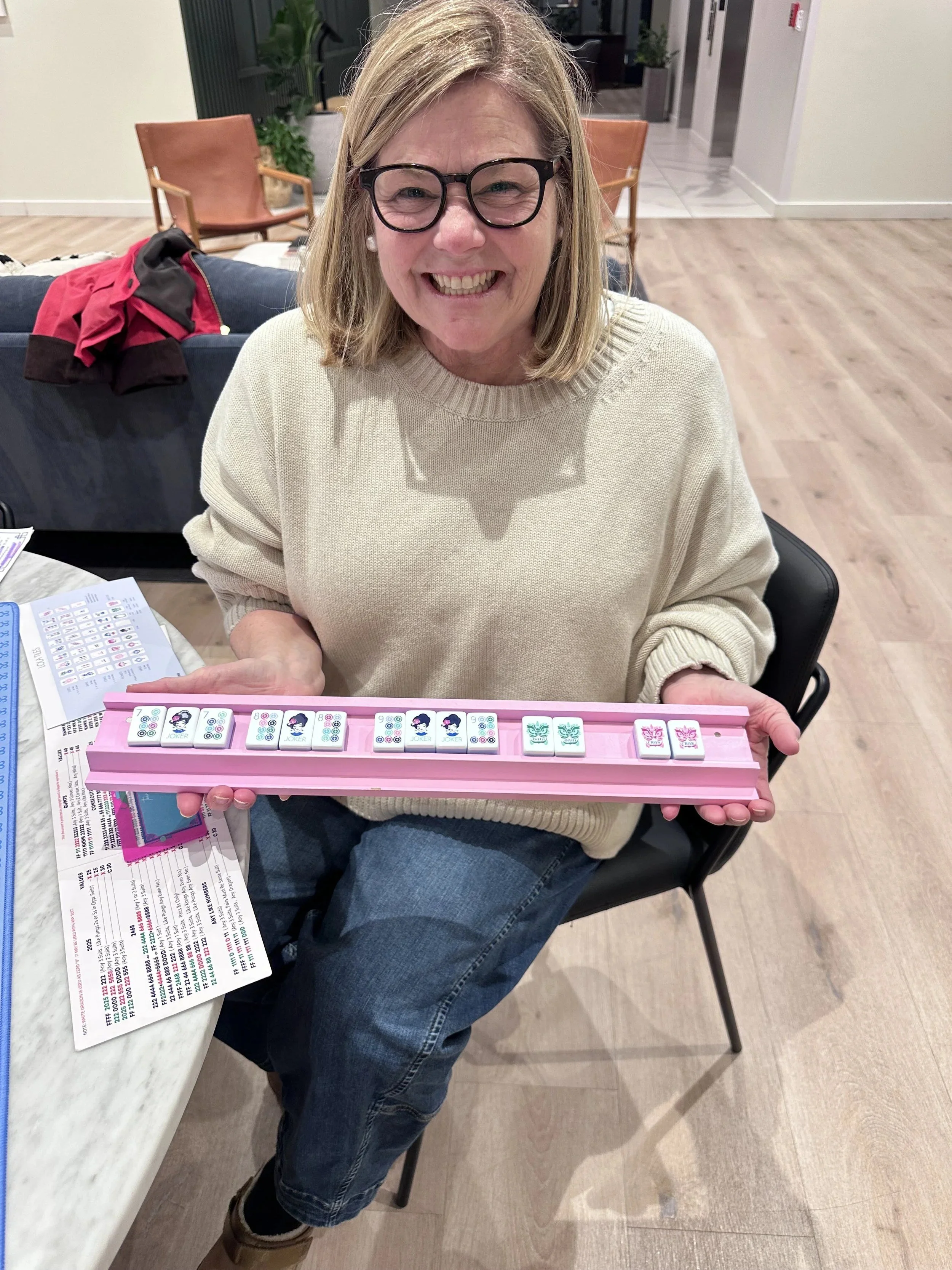 A woman wearing glasses and a beige sweater, smiling, holding a pink Mahjong tile rack with Mahjong tiles, sitting at a table with lottery tickets and a card in front of her in an indoor setting.