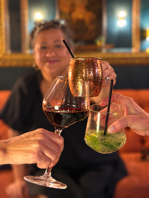 Three people holding different glasses: a red wine glass, a copper mug, and a glass with a green cocktail, in a dimly lit restaurant.
