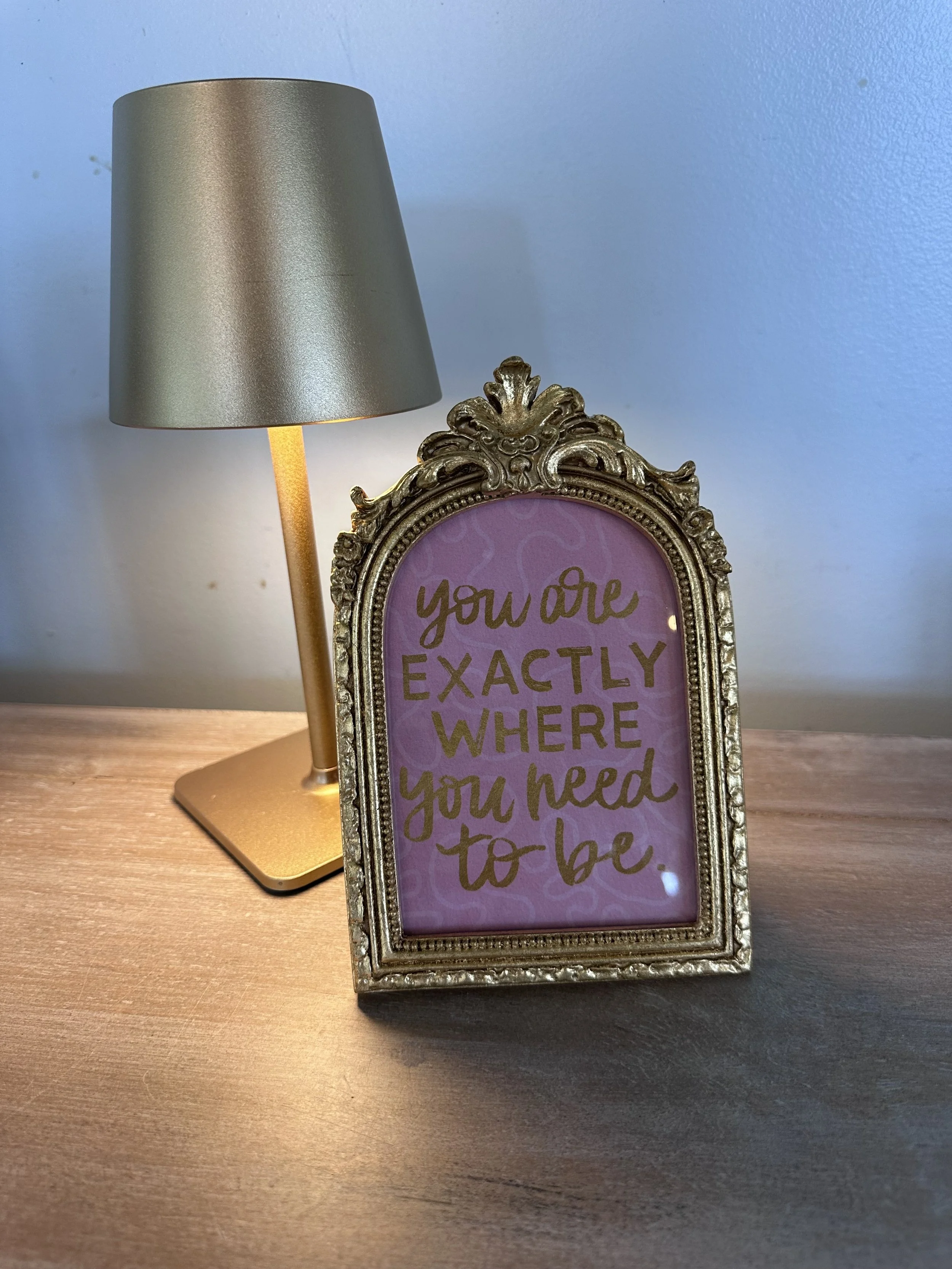 A gold and black table lamp and a gold ornate picture frame with a pink background and gold lettering that says "You are exactly where you need to be."