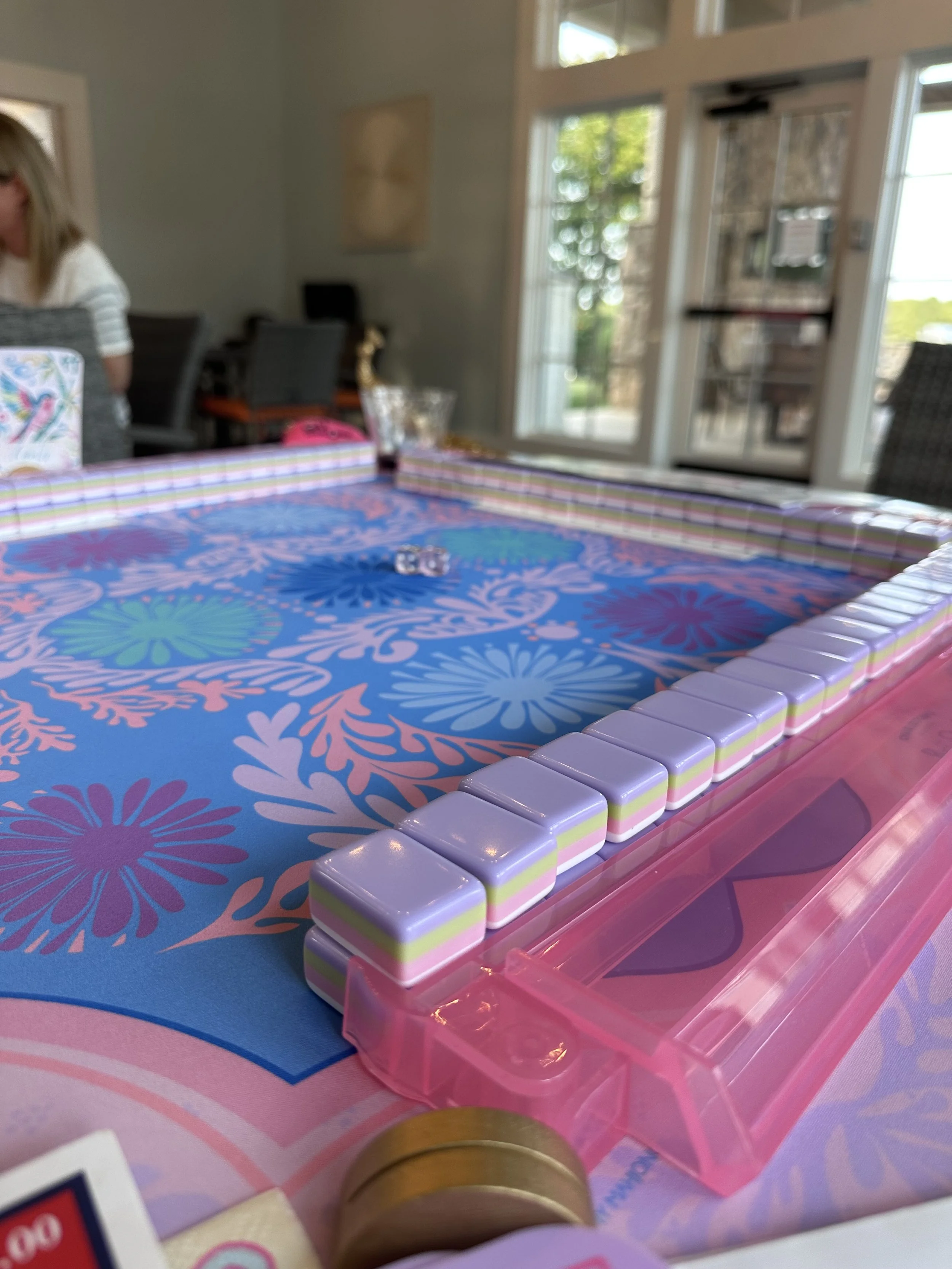 Close-up of a colorful tabletop game with a blue and pink floral design, surrounded by striped edges and a pink plastic container nearby. In the background, a woman with blonde hair is partially visible, sitting at a table in a well-lit room with lar