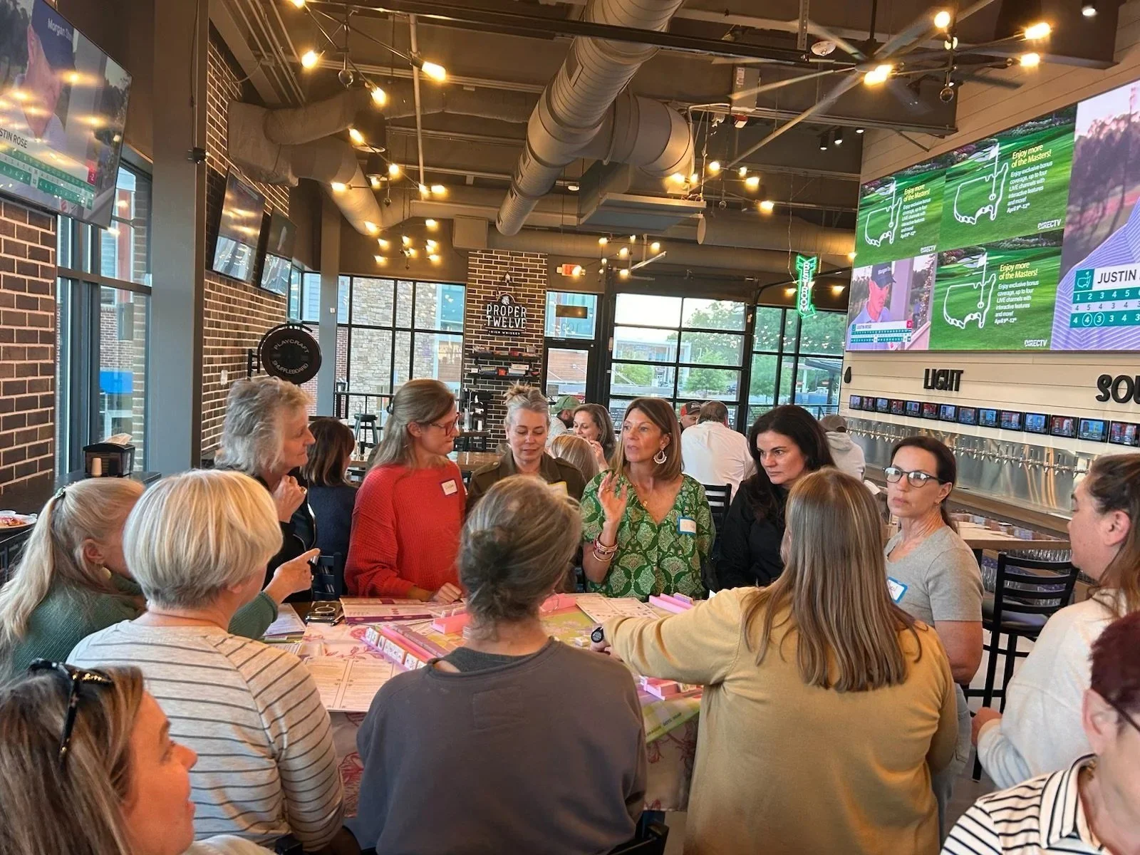 A group of women gathered around a table in a restaurant, engaging in conversation. The interior features large windows, brick walls, and bright overhead lighting, with a large digital screen displaying a golf game in the background.
