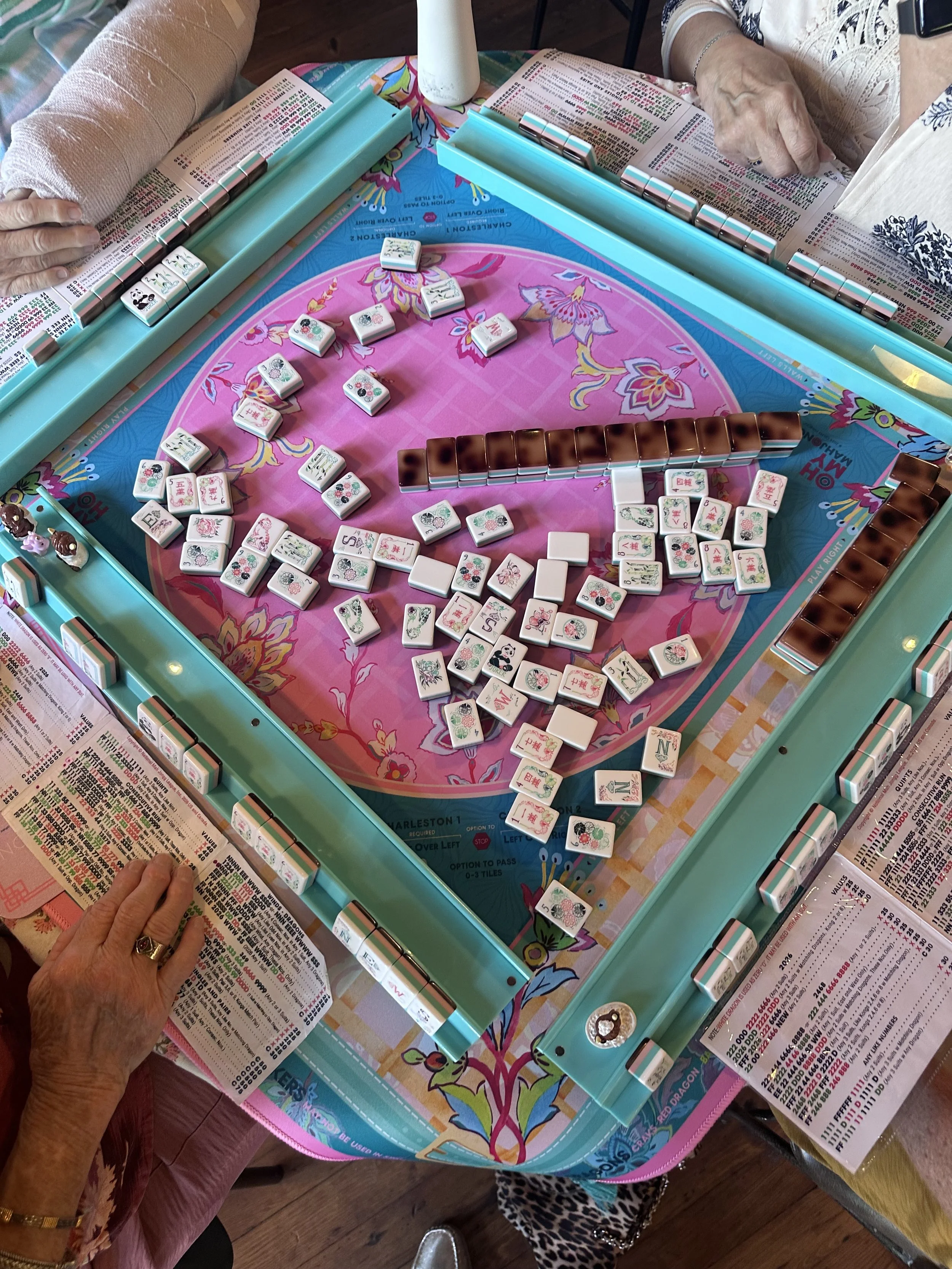 A close-up view of a Mahjong game in progress on a brightly colored table, with two players, one of whom has a visible hand and wrist. The table features tiles with traditional Chinese characters and symbols, and game instruction papers are seen arou