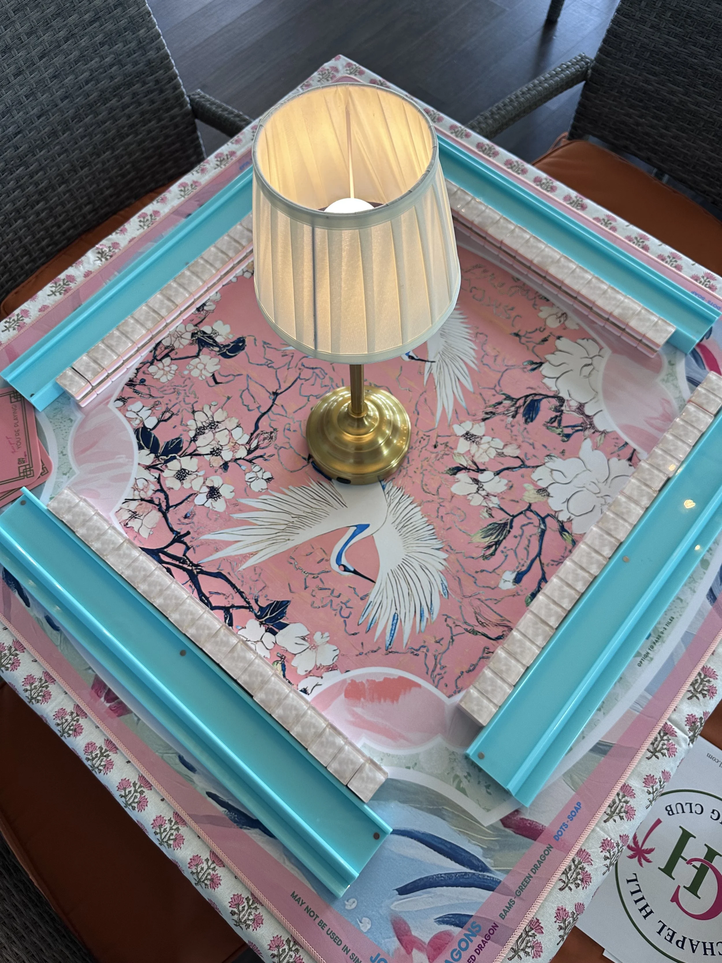 A decorative table with a pink background featuring white sakura blooms and a white crane. On the table, there is a brass table lamp with a cream-colored lampshade, and plastic strips in mint green, pink, and light blue along the border. The table is