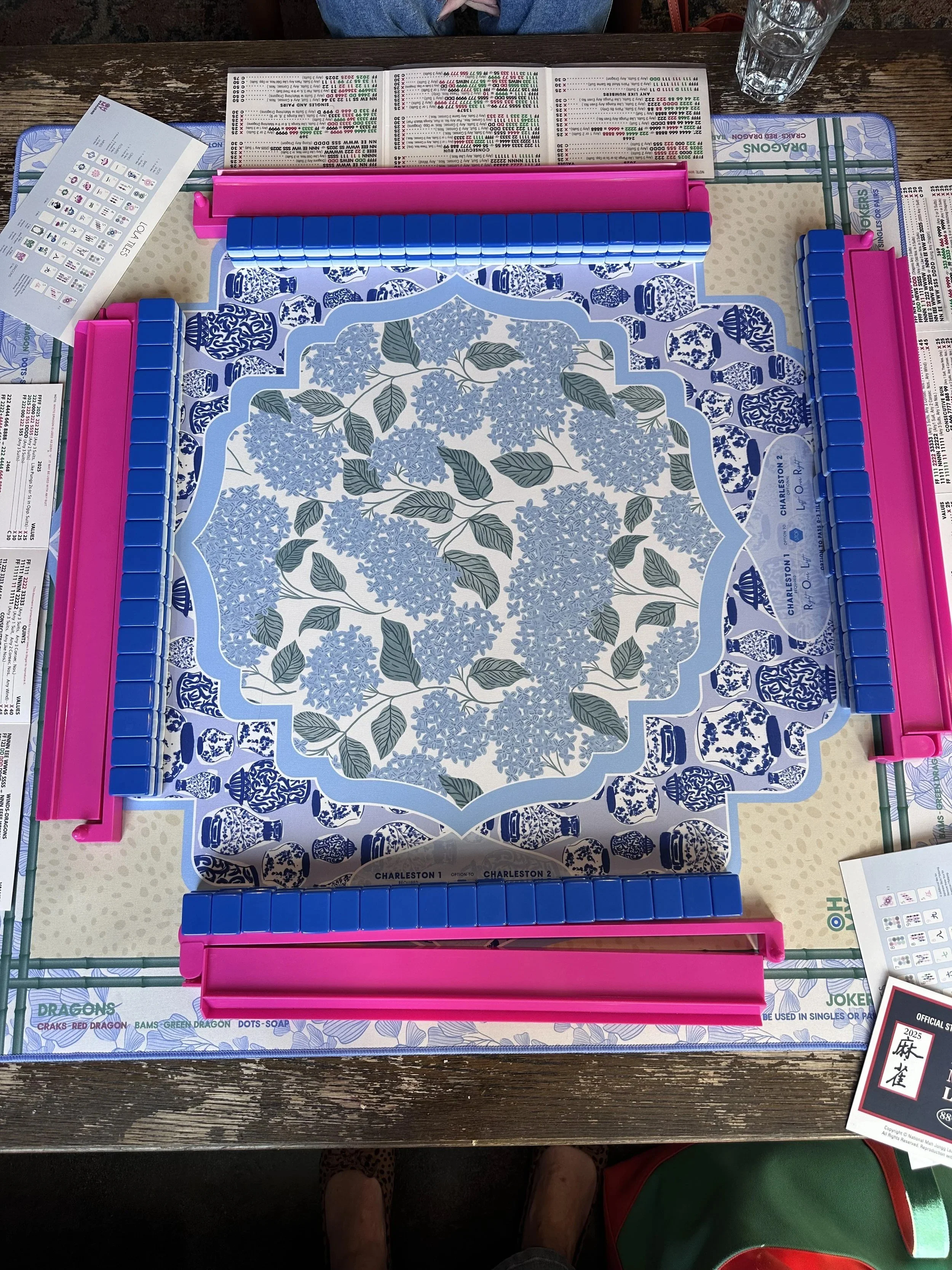 A Bananagrams game setup with a patterned tile rack, some game tiles, and rule sheets on a wooden table, with a glass of water and a person wearing shorts and sandals sitting nearby.