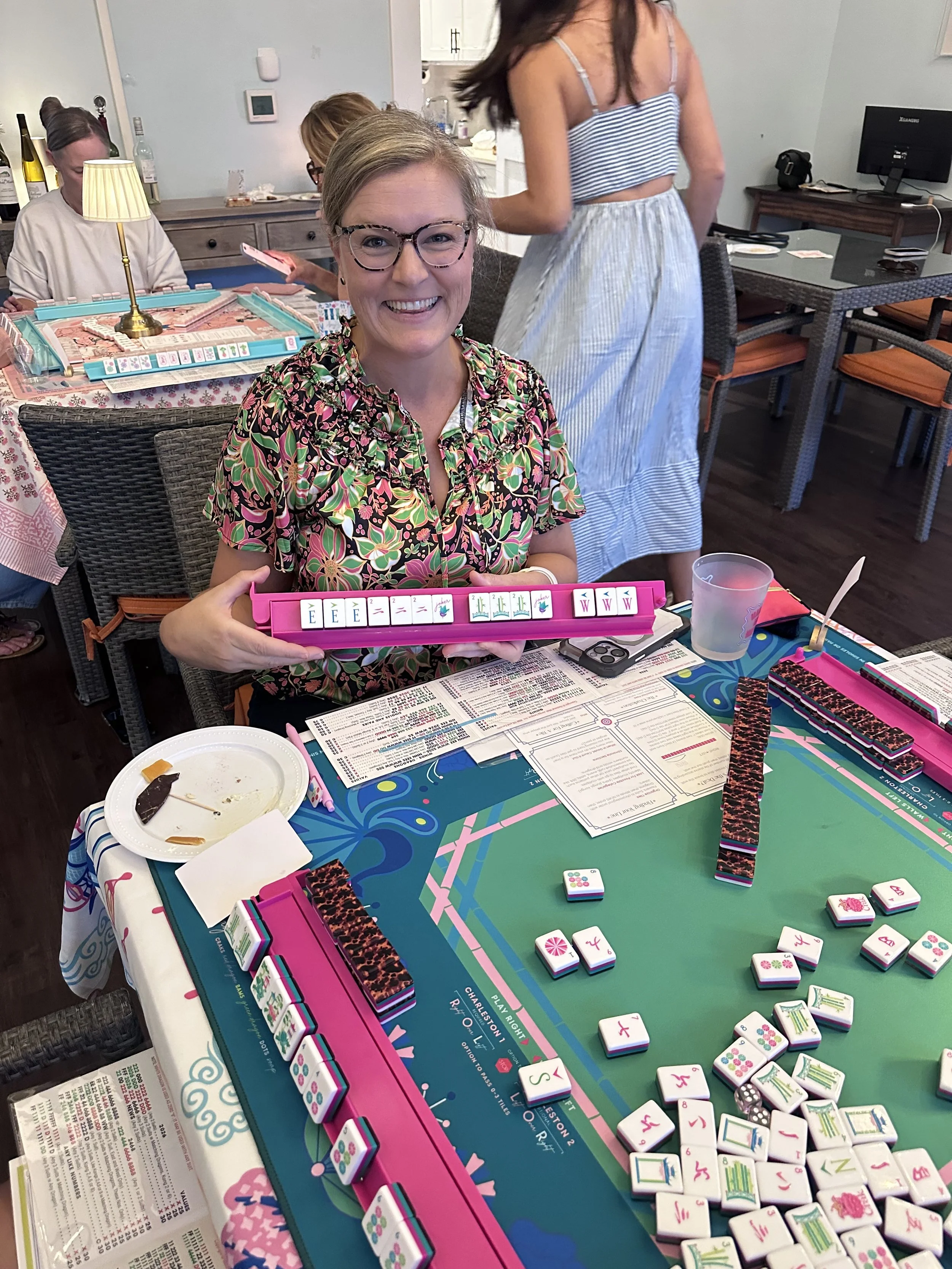 A woman smiling and holding a set of colorful Mahjong tiles at a table with Mahjong game pieces, rule sheets, and snacks in a restaurant or cafe setting.