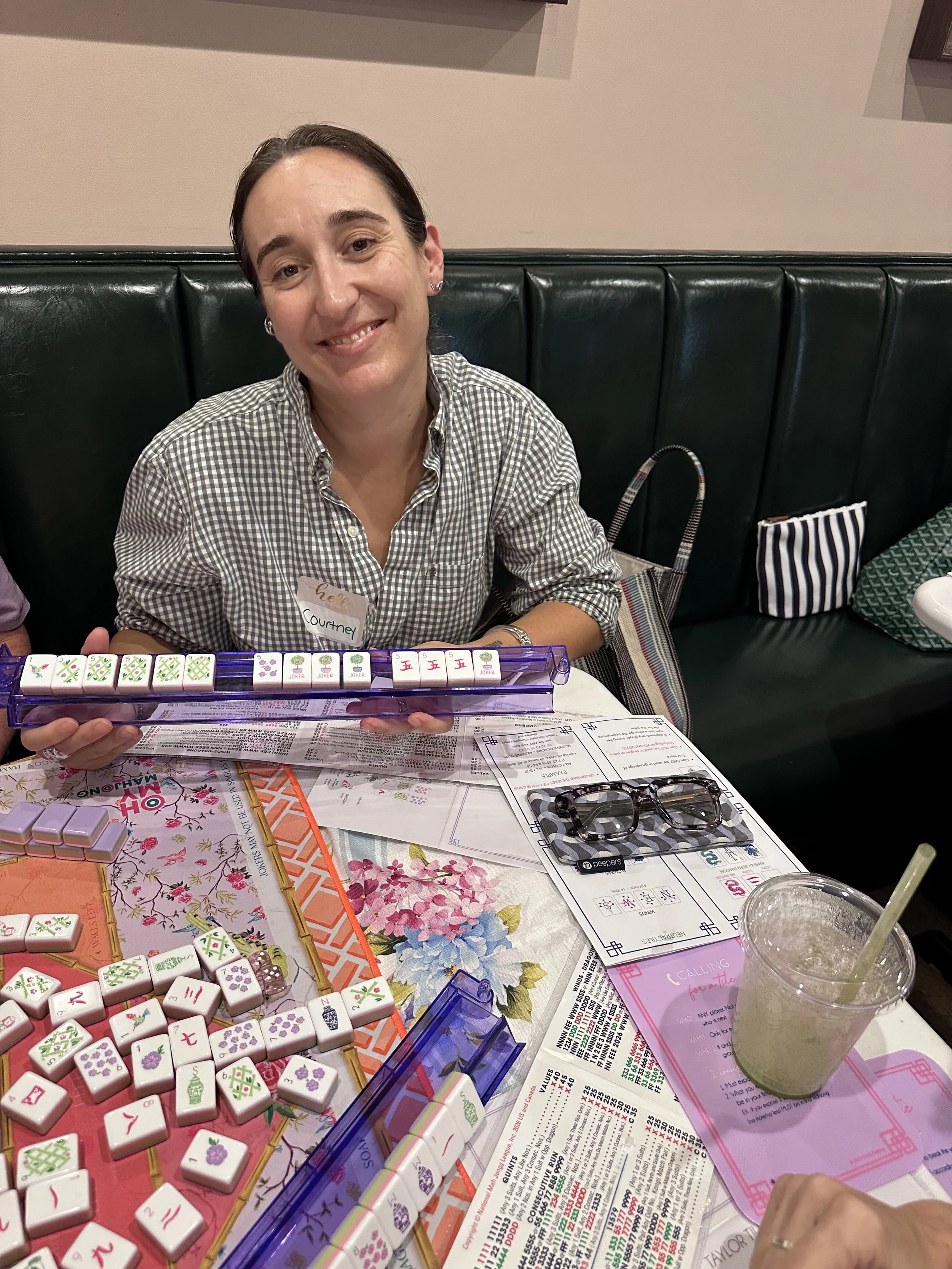 A woman sitting at a table playing mahjong, surrounded by mahjong tiles, papers, and a drink with a straw.