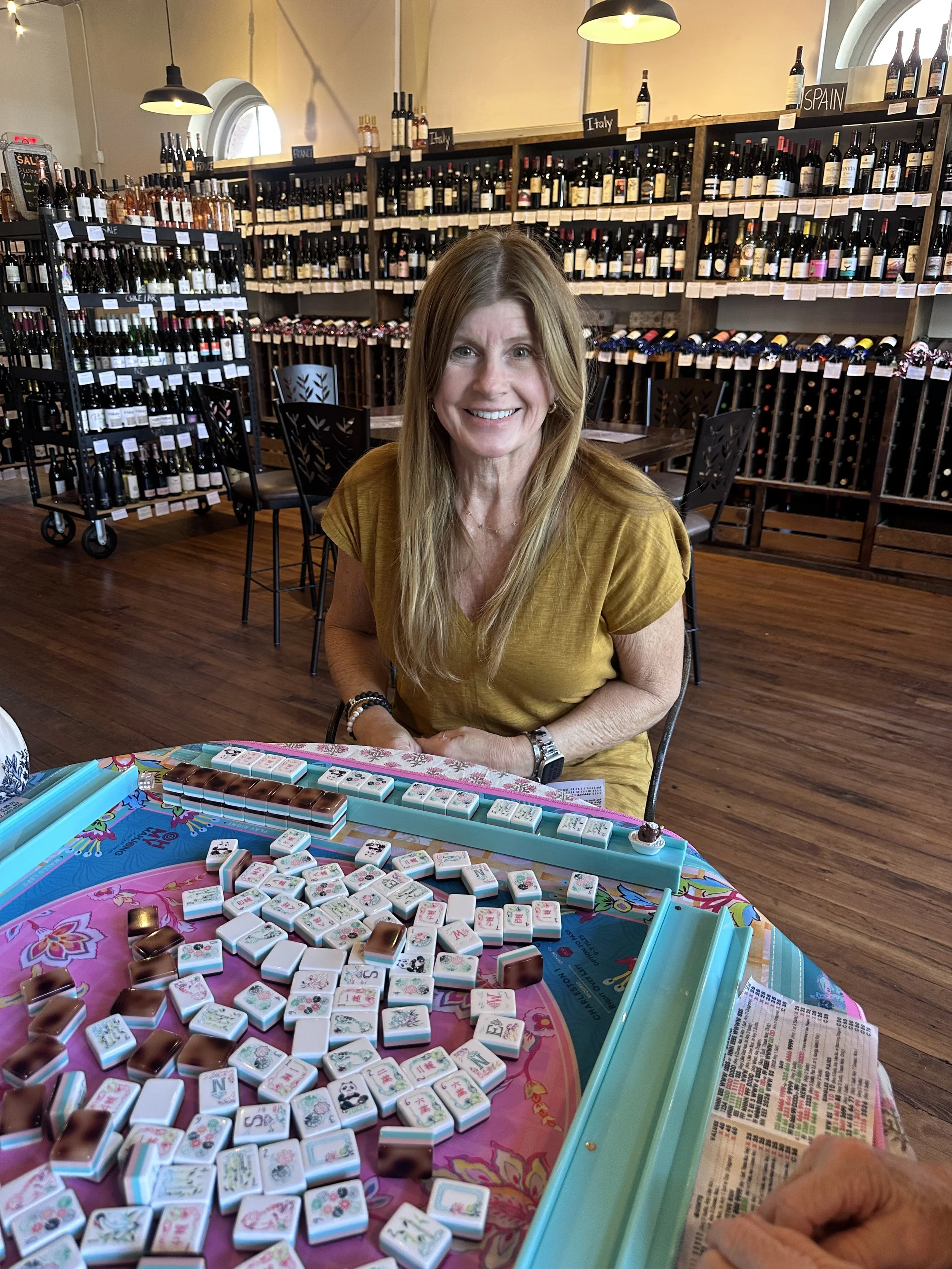 A woman sitting at a table playing Mahjong, with a colorful Mahjong game set in front of her. The background shows shelves with wine bottles labeled by country, and a window with natural light.