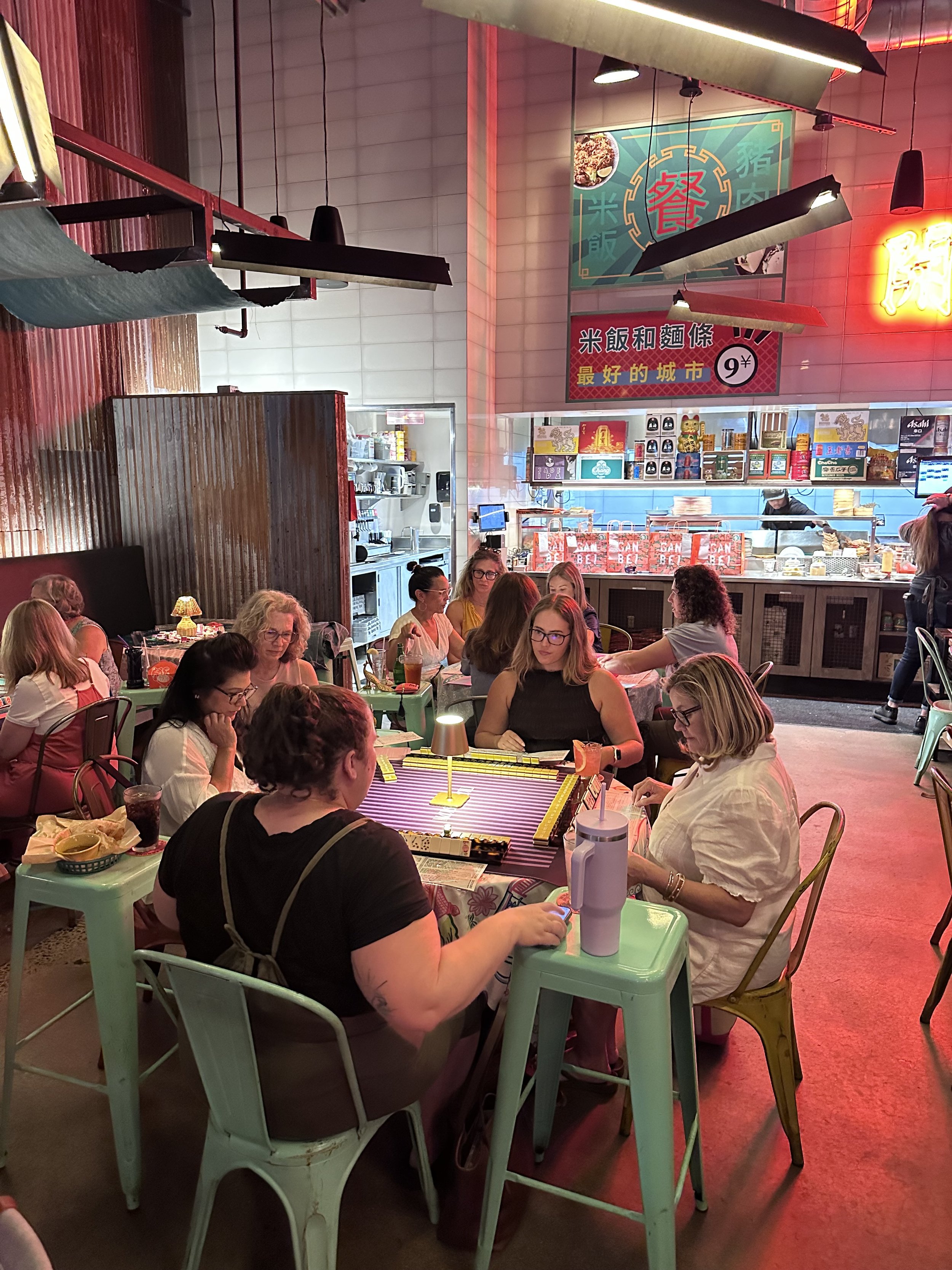 People dining in a Chinese restaurant, with a group of women sitting at tables, some looking at menus, and a kitchen area visible in the background. The restaurant has neon signs and Asian decor.