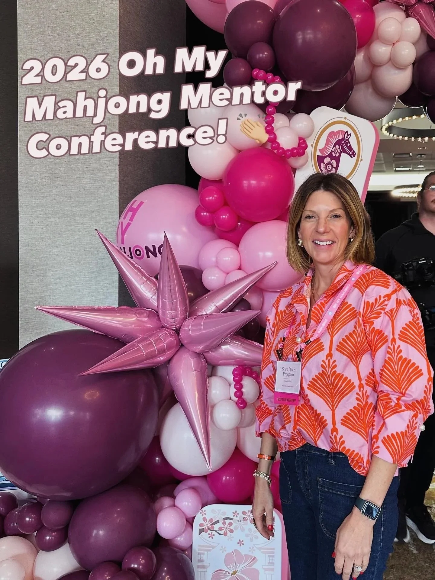 A woman standing in front of a pink and purple balloon display at the 2026 Oh My Mahjong Mentor Conference, with festive balloons and a sign with a pink horse logo.