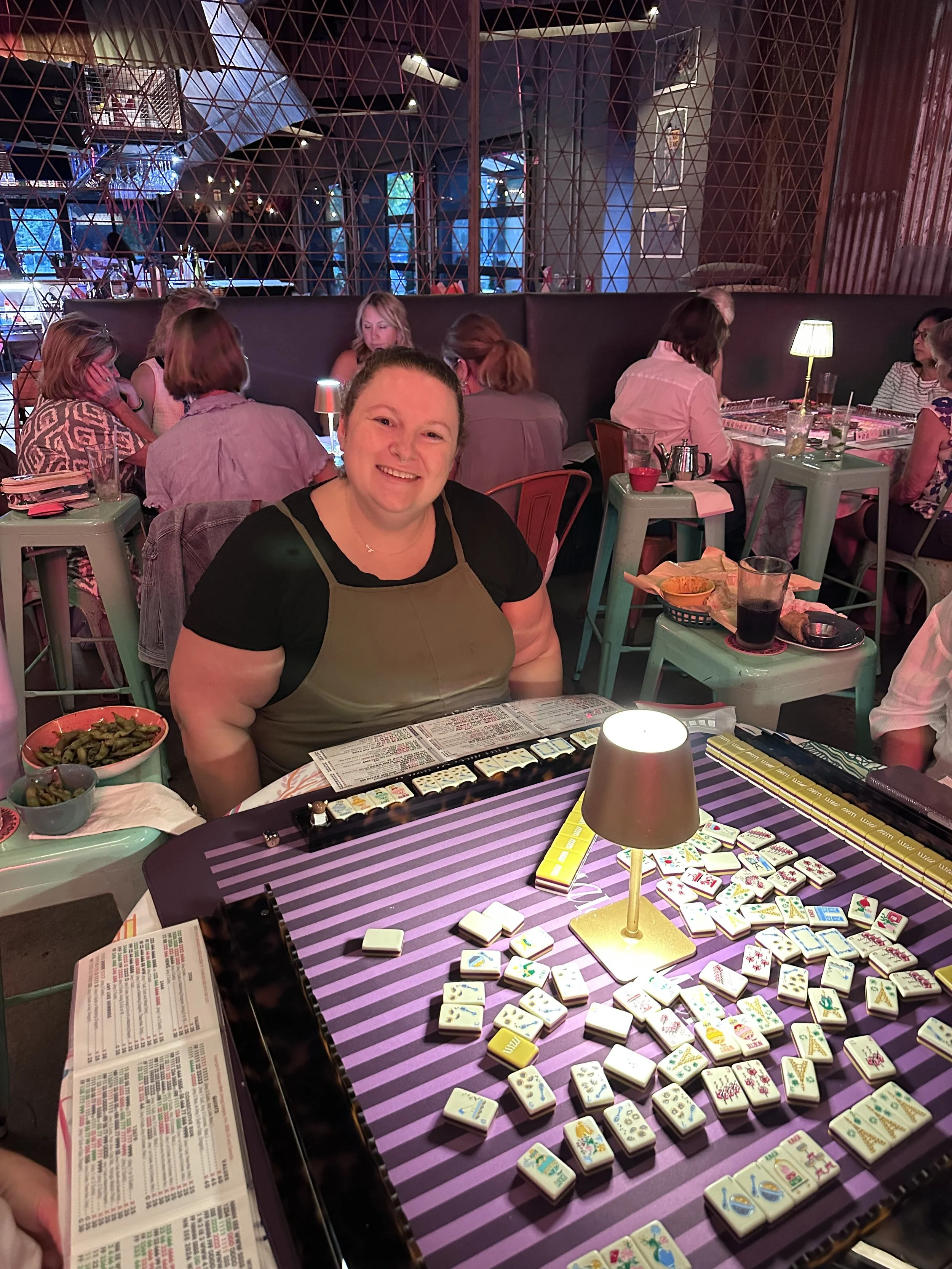 A woman sitting at a table with mahjong tiles and score sheets in a restaurant or bar setting. Other people are seated at tables in the background, dining and socializing. The room has a modern, cozy decor with dim lighting and decorative framed pict