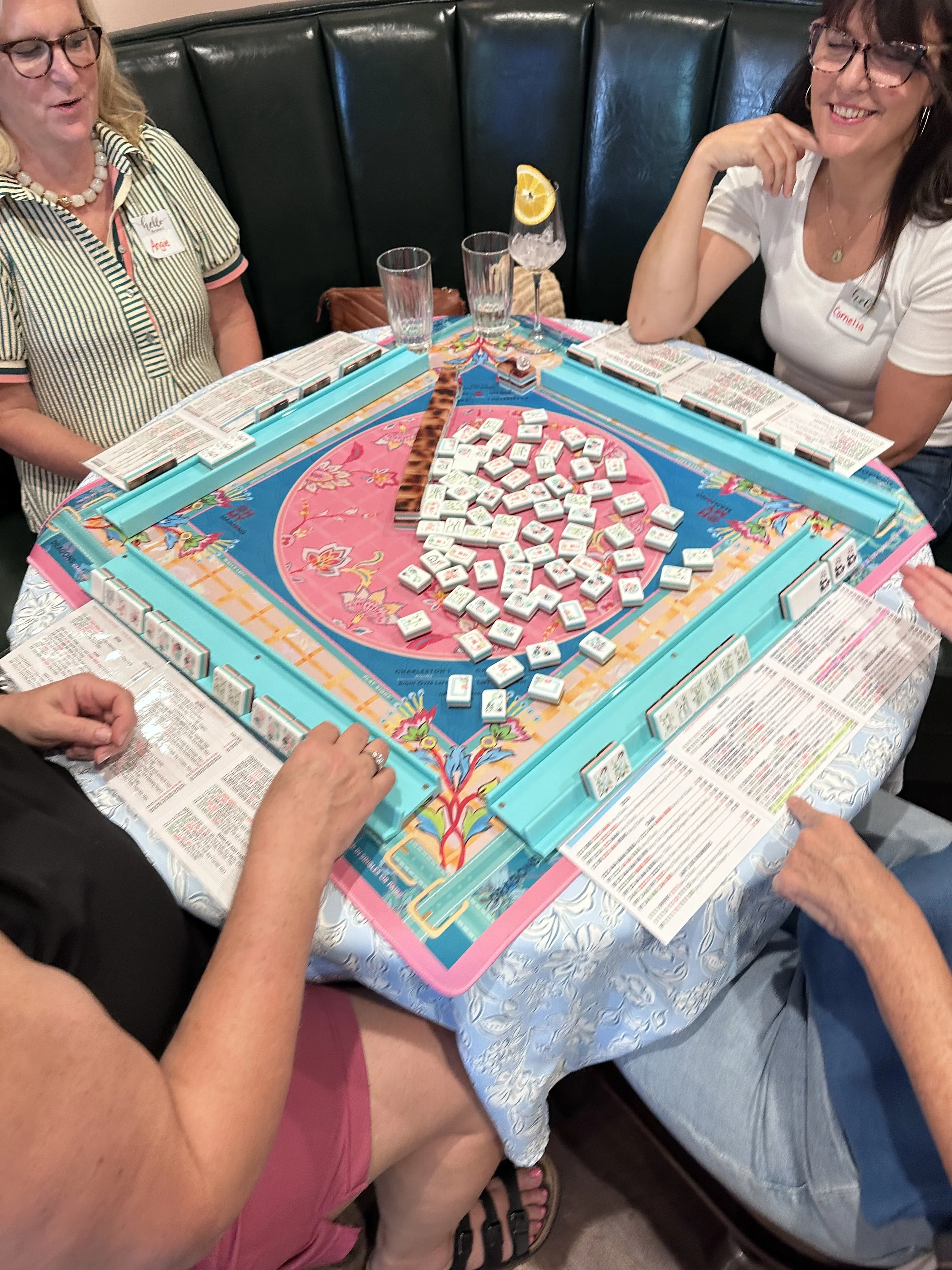 People playing a game of Mahjong at a round table with a colorful tablecloth, with drinks including a cocktail with an orange slice. The players are women, with glasses and casual clothing, engaged in the game.