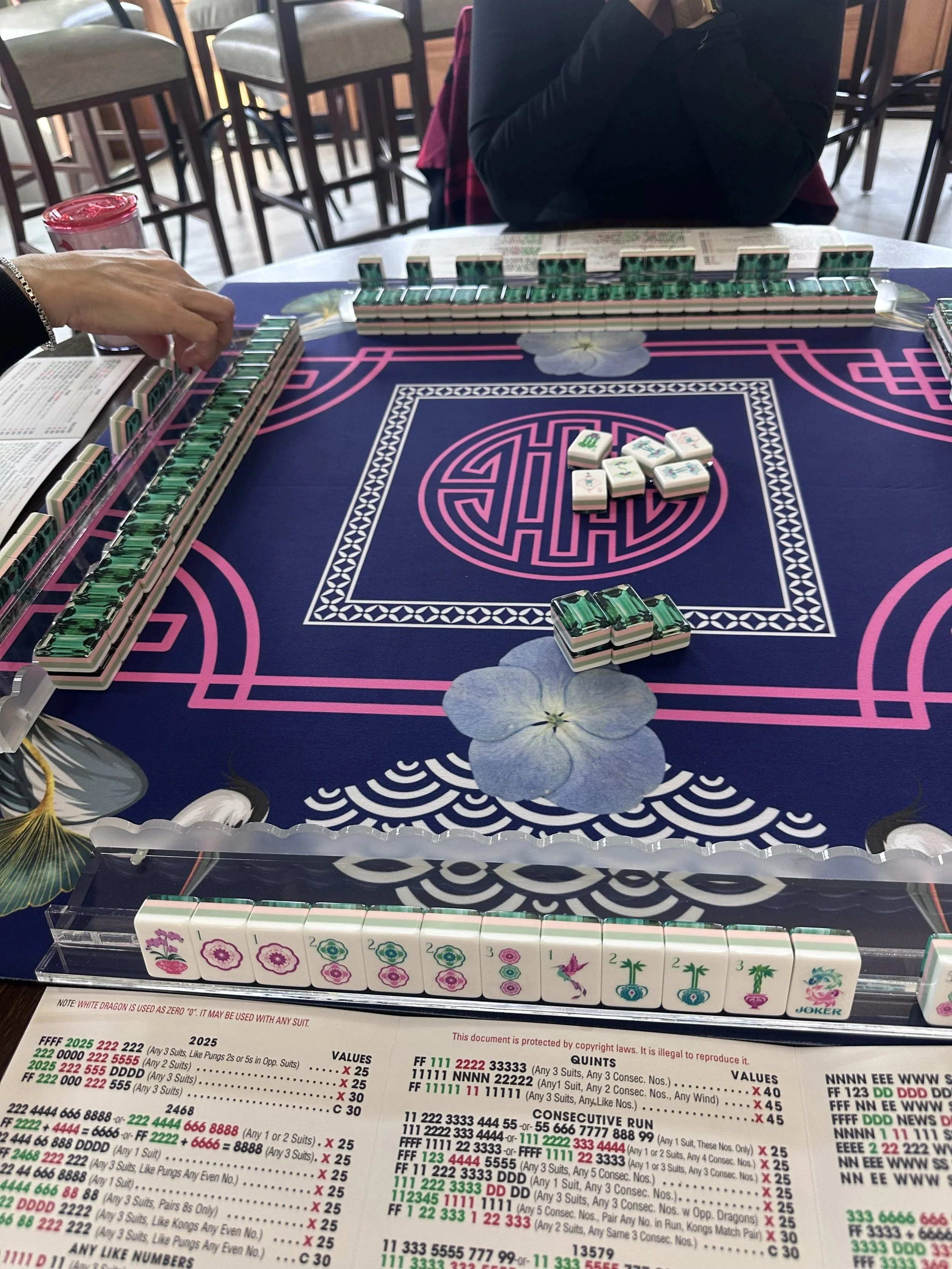 Close-up of a Mahjong game in progress on a table, with tiles arranged in front and a player's hand reaching to pick a tile. In the background, a person with crossed arms is seated, and other chairs and tables are visible in a casual dining or game r