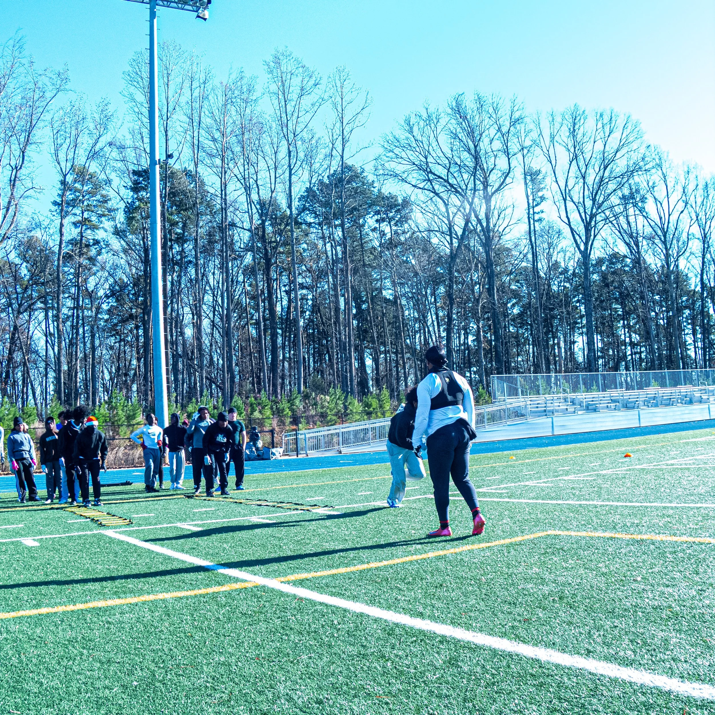A group of people on a sports field, some standing in line and others walking around, with trees and clear blue sky in the background.