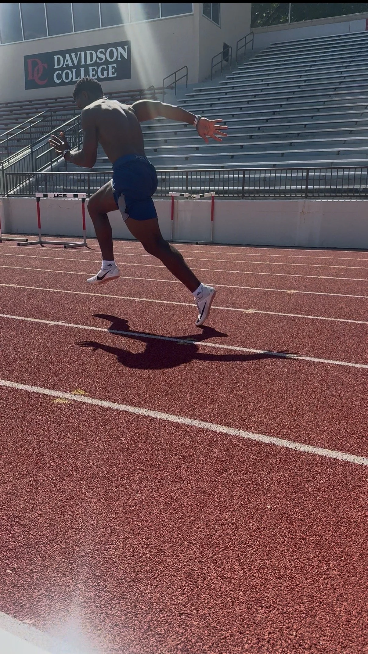 A male athlete in blue shorts and white sneakers sprinting on an outdoor track at Davidson College, with empty bleachers and a building in the background.