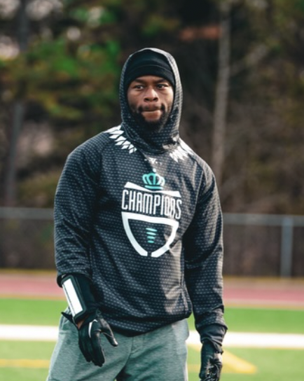 A young man dressed in athletic clothing standing outdoors on a sports field, wearing a hoodie, gloves, and a bandana around his face.